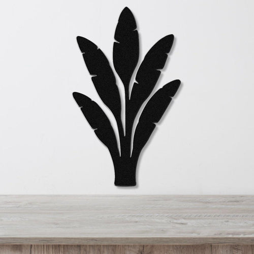 Black metal wall art in a banana leaf silhouette with layered cutouts, mounted on a white wall above a light wood counter.