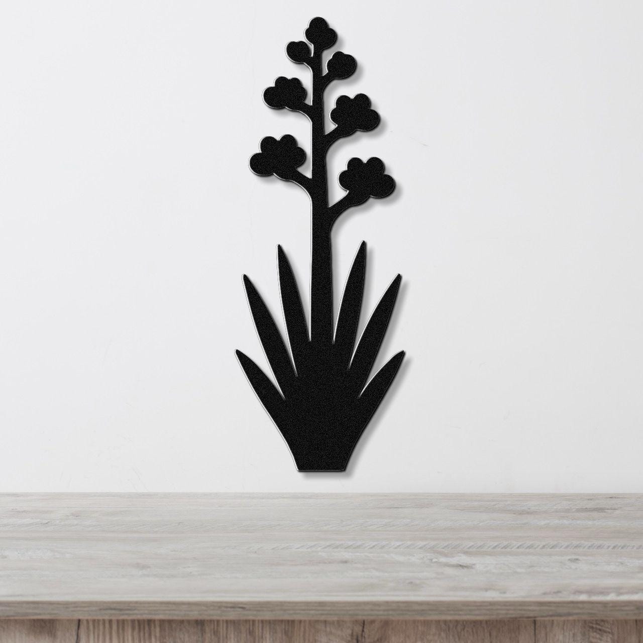 Black metal wall art silhouette of a blooming agave plant with spiky leaves and flower clusters, mounted on a white wall above a light wood surface.