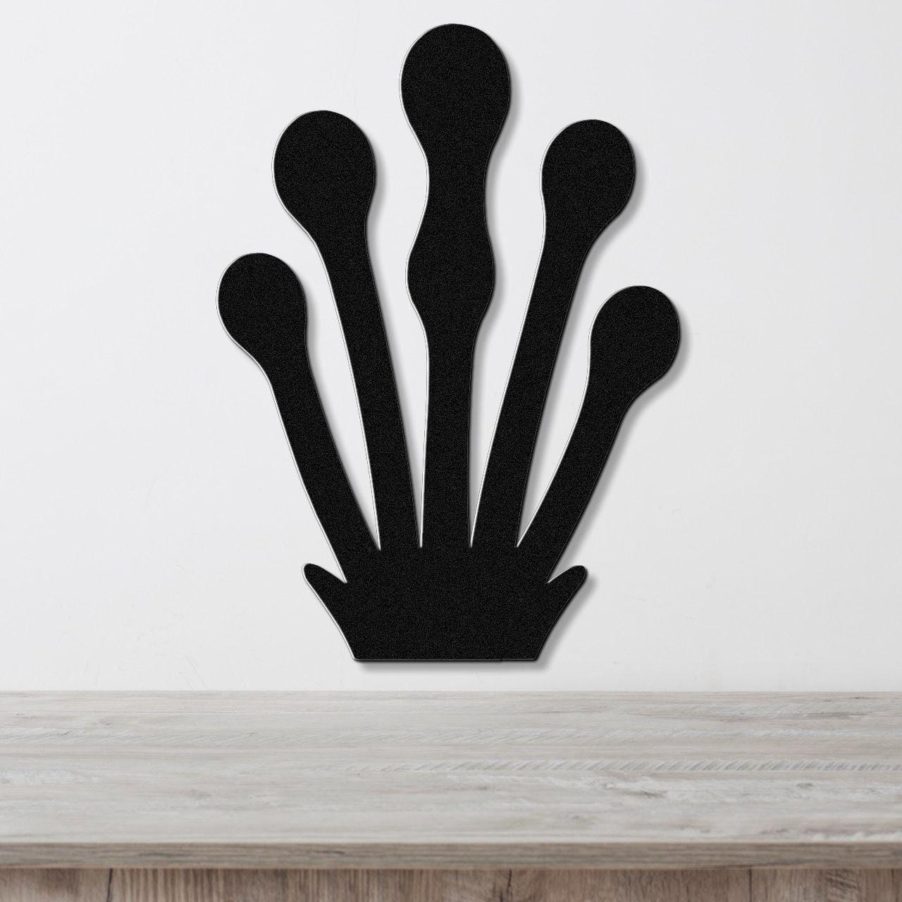 Black metal wall art in a kelp or seaweed silhouette design with five elongated fronds, mounted on a white wall above a light wood surface.