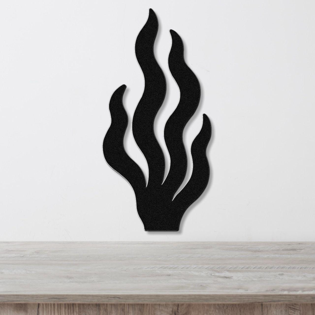 Black metal wall art in a kelp or seaweed silhouette with layered, flowing fronds, mounted on a white wall above a light wood surface.