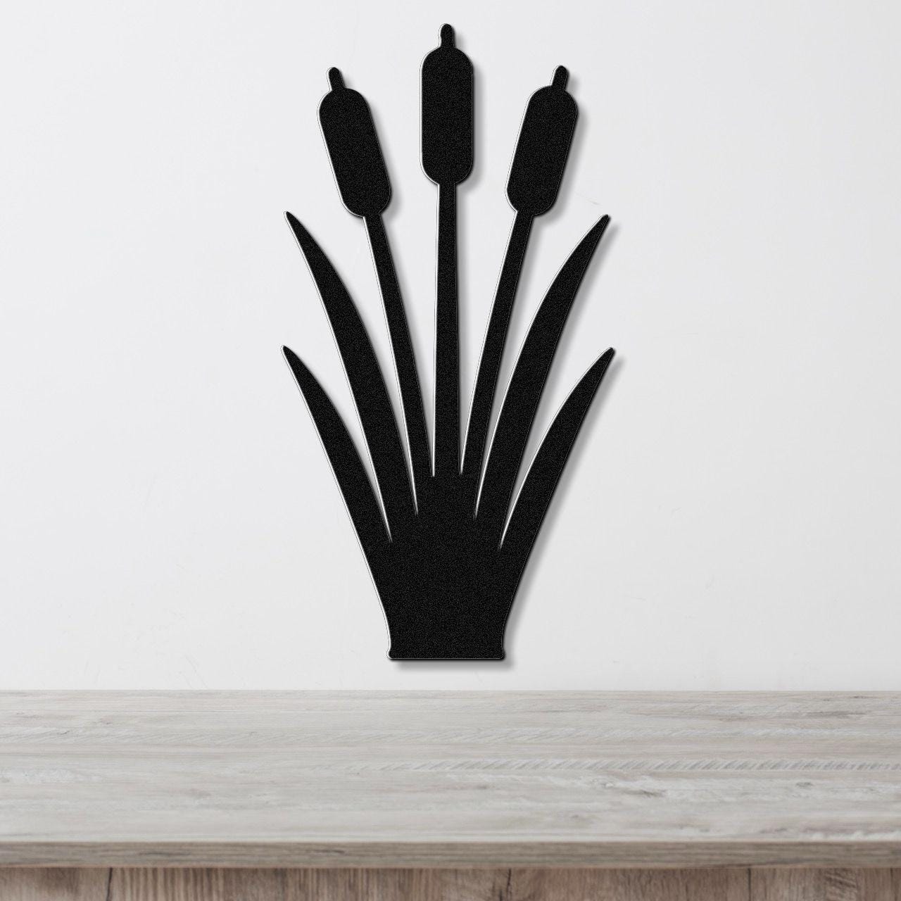Black metal cattail wall art silhouette with three seed heads and long leaves, mounted on a white wall above a light wood surface.