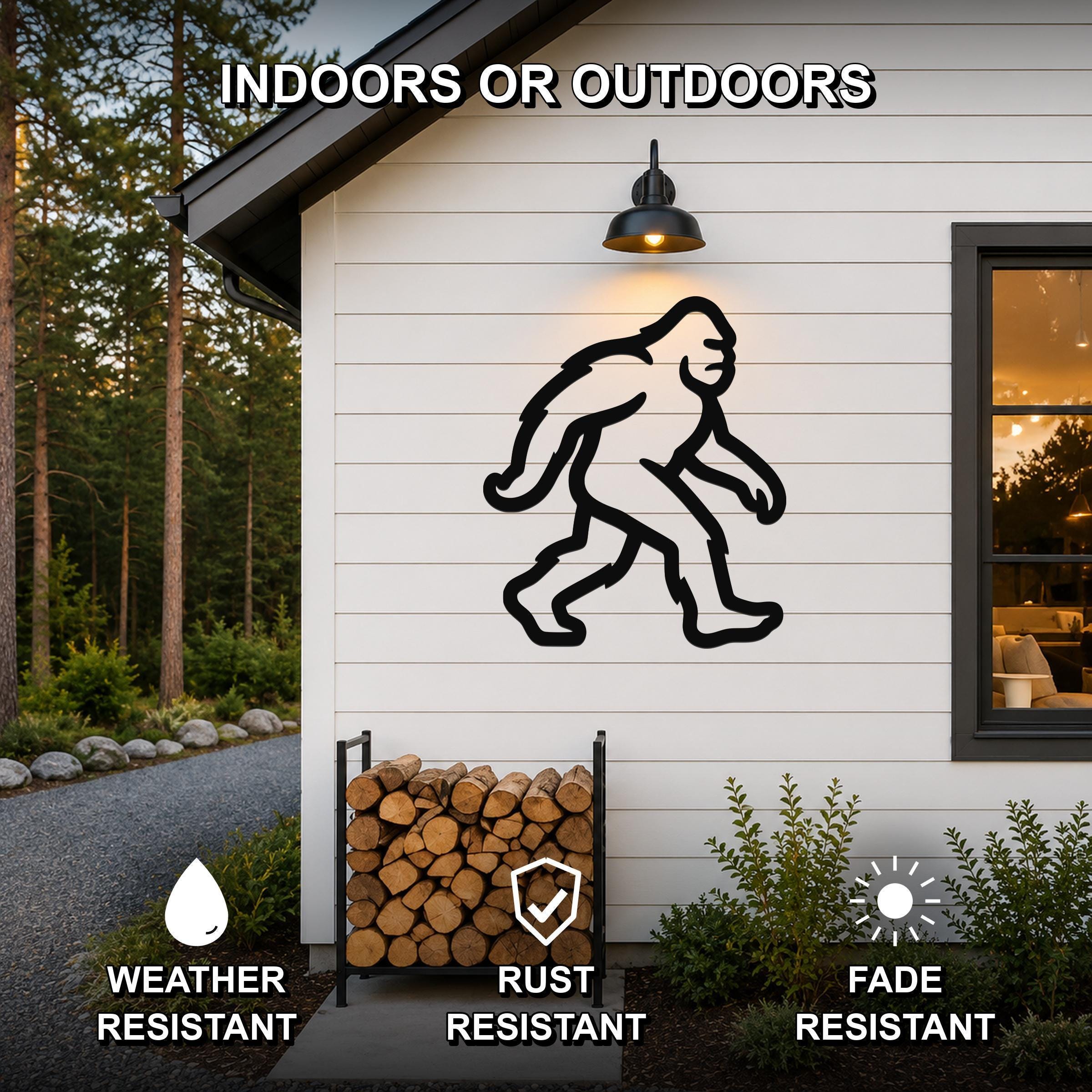 Black metal wall art featuring a Bigfoot silhouette, mounted on a white exterior wall above a firewood rack, with text indicating it is weather, rust, and fade resistant for indoor or outdoor use.