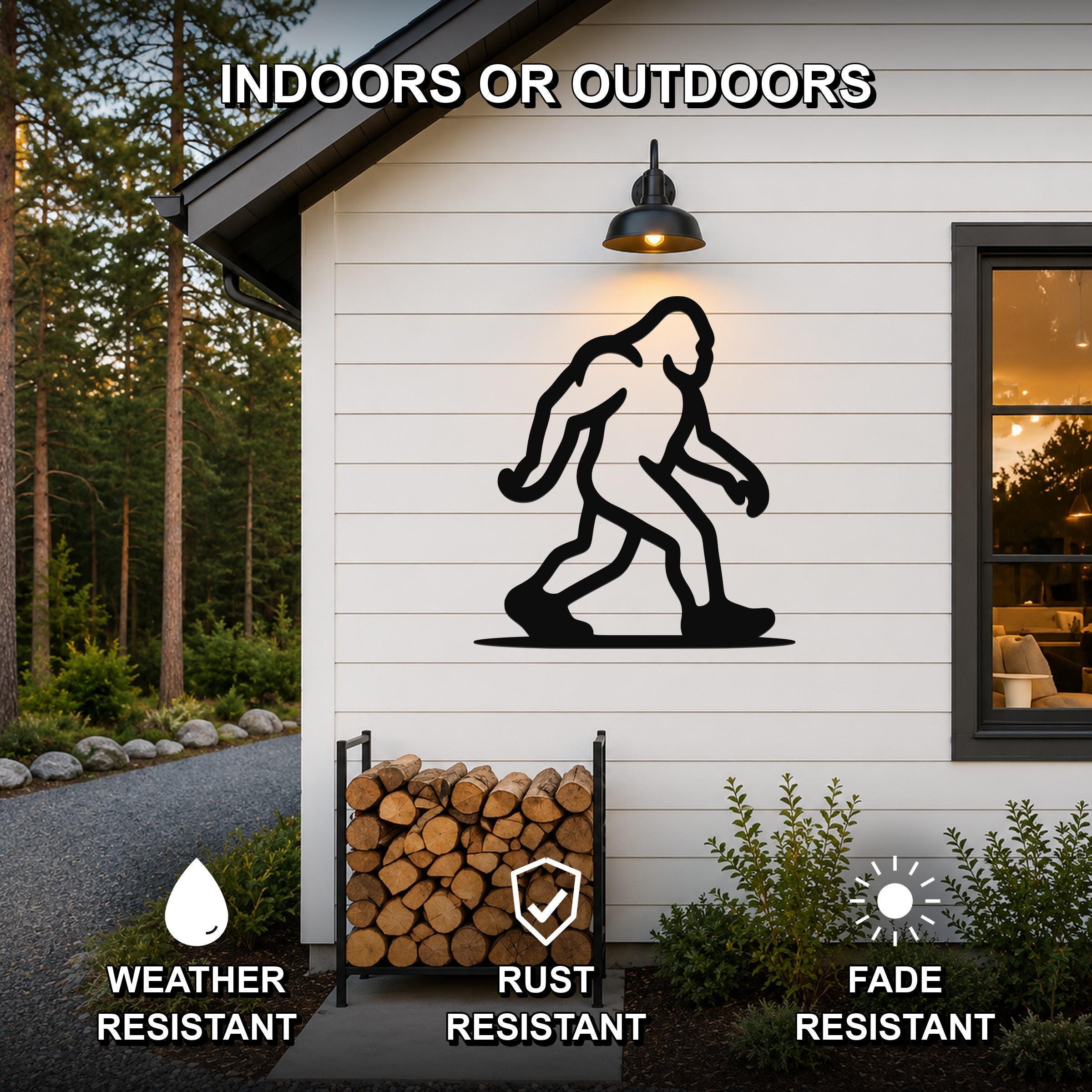 Black metal Bigfoot silhouette wall art mounted on a white siding exterior wall above a firewood rack, with a lit outdoor sconce above and a window to the right.