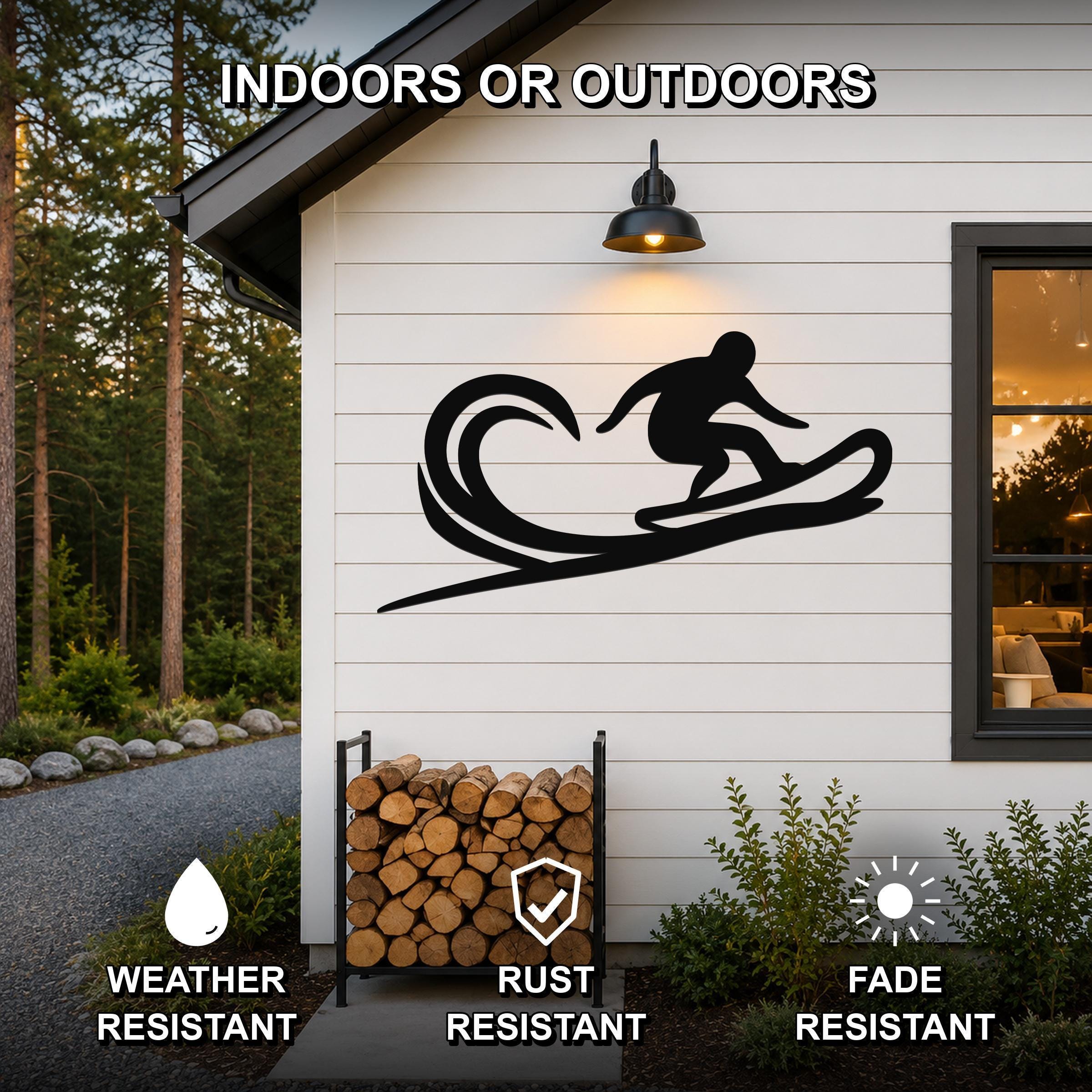 Black metal wall art featuring a surfer silhouette on a wave, mounted on a white exterior wall above a firewood rack, with text indicating it is weather, rust, and fade resistant for indoor or outdoor use.