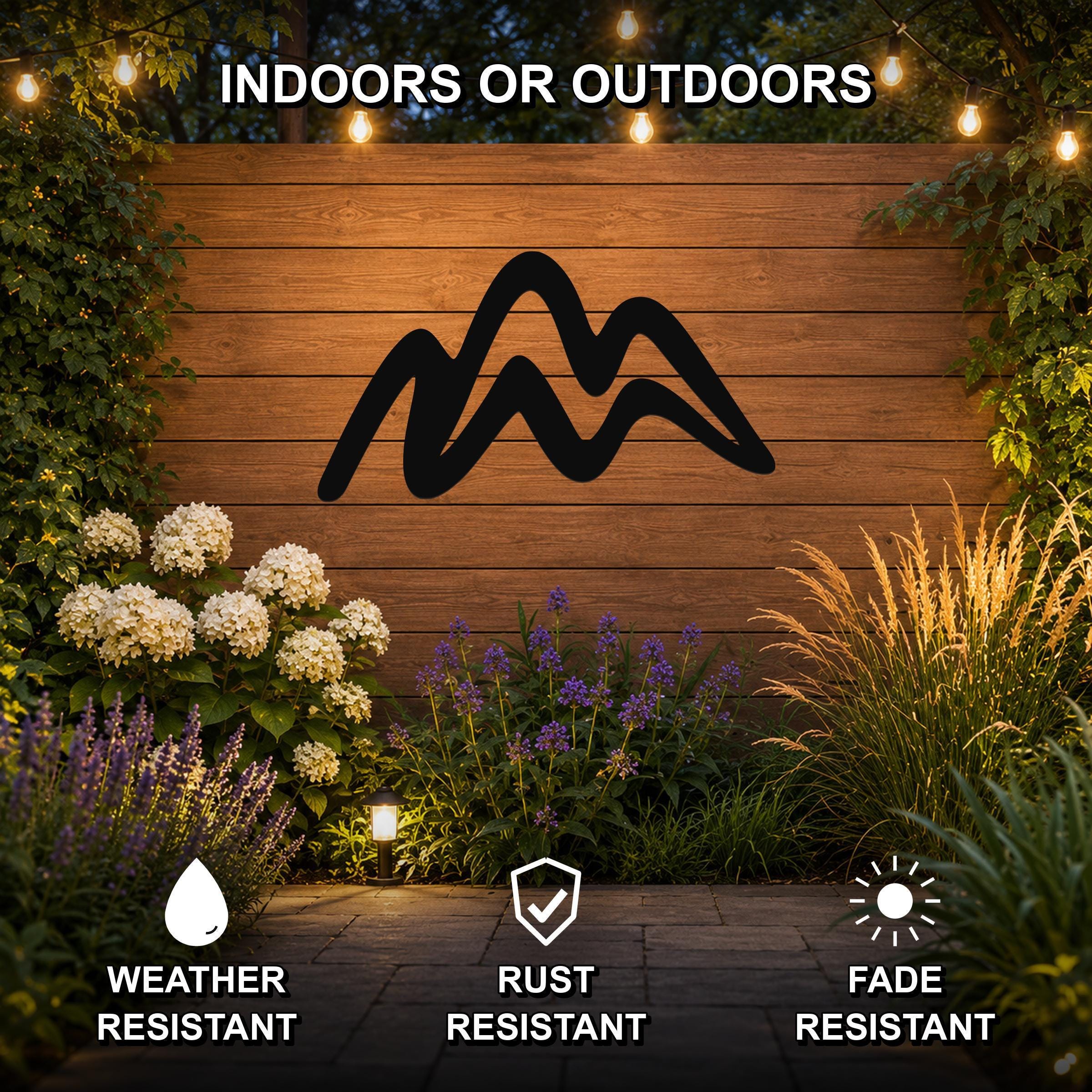 Black metal mountain silhouette wall art mounted on a wooden outdoor wall, surrounded by plants and string lights, with text indicating it is weather, rust, and fade resistant.