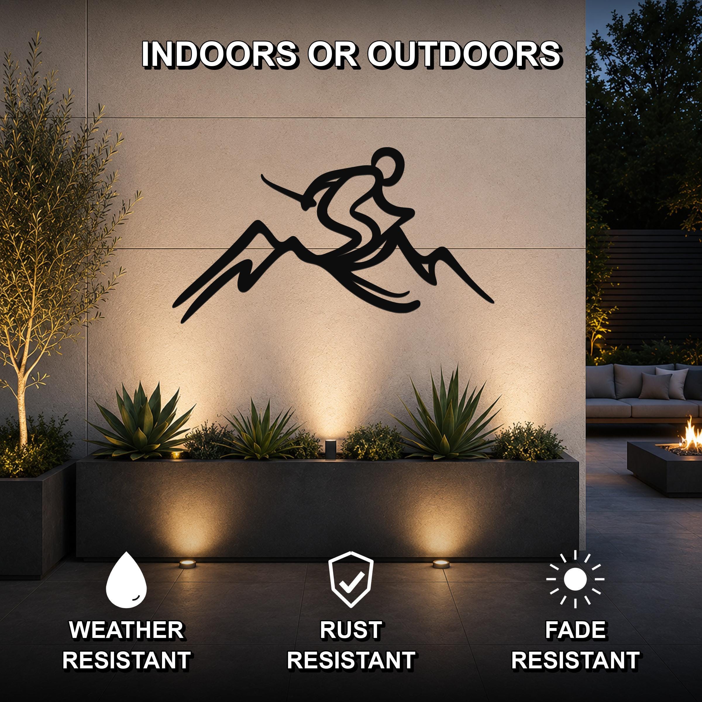 Black metal skier and mountain wall sculpture with dynamic flow cutouts, mounted on an outdoor patio wall above a planter with uplighting.