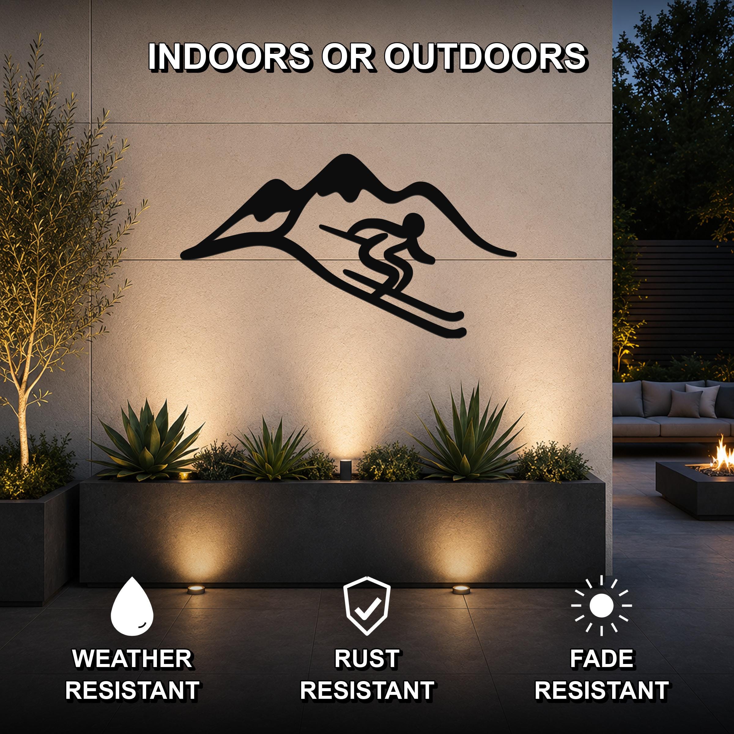 Black metal skier and mountain wall art silhouette mounted on an outdoor patio wall above a planter, with text indicating it is weather, rust, and fade resistant.