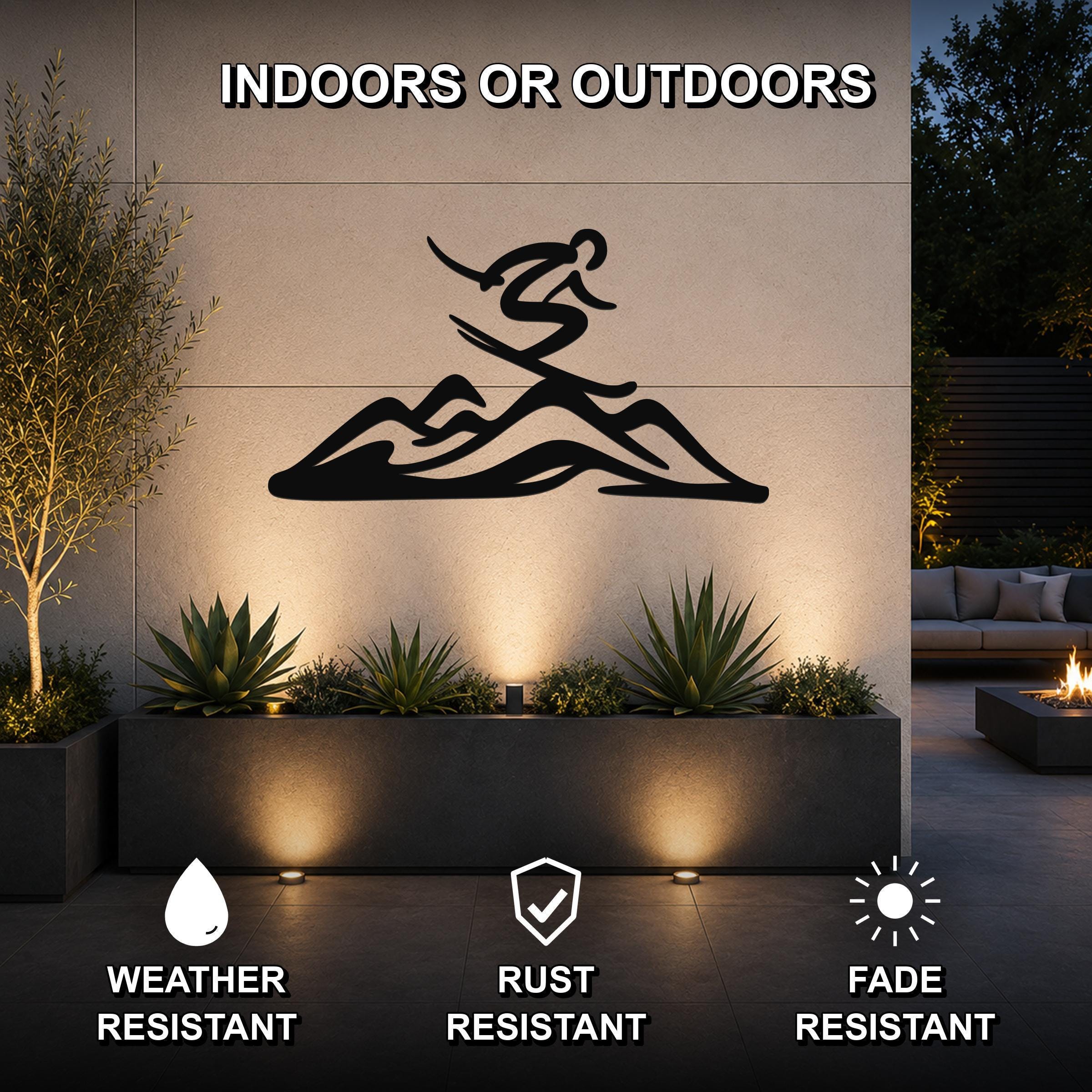 Black metal skier and mountain wall art silhouette mounted on an outdoor patio wall above a planter, with text indicating it is weather, rust, and fade resistant.
