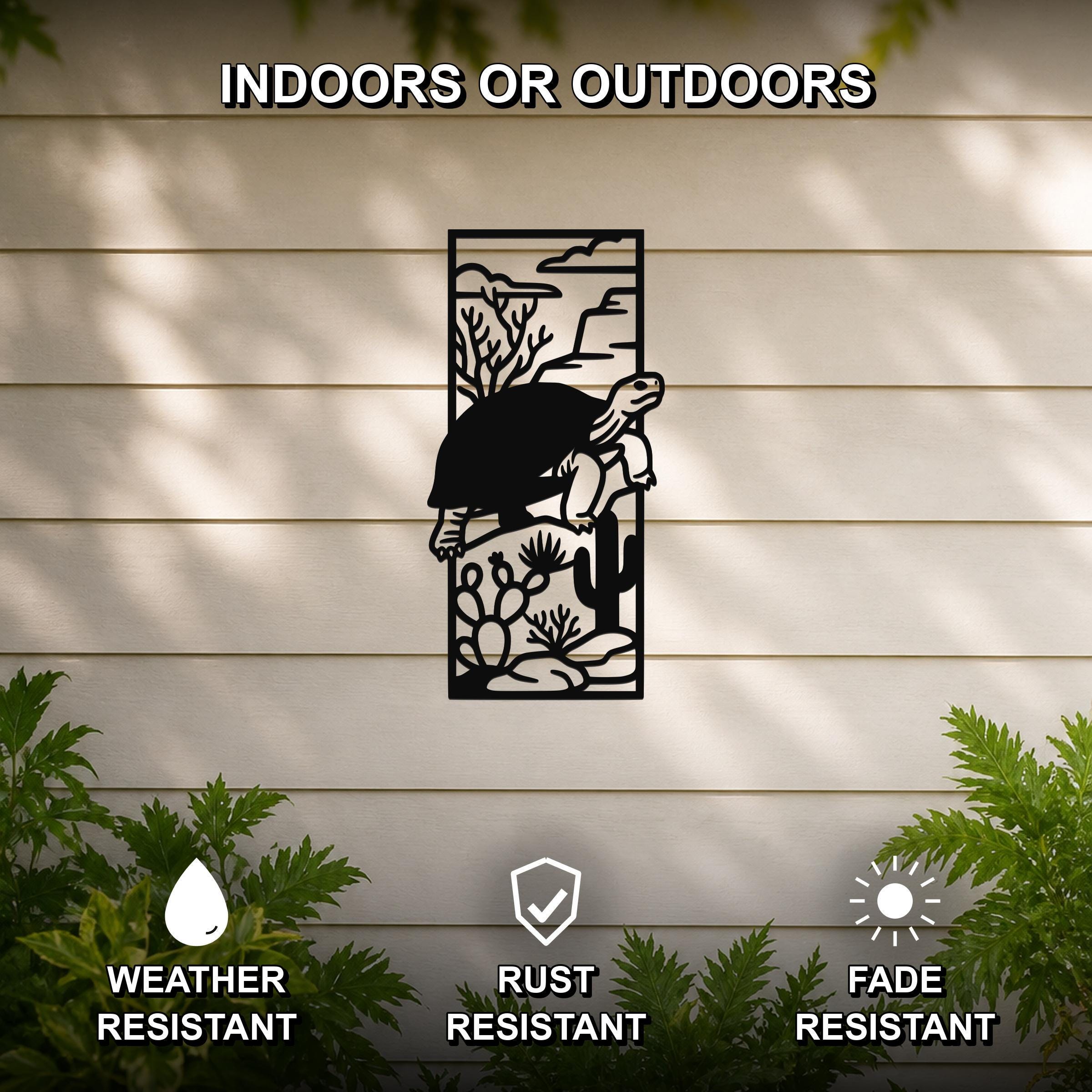 Black metal wall art featuring a tortoise silhouette climbing over cacti and desert plants, mounted on a light-colored outdoor wall with greenery below.