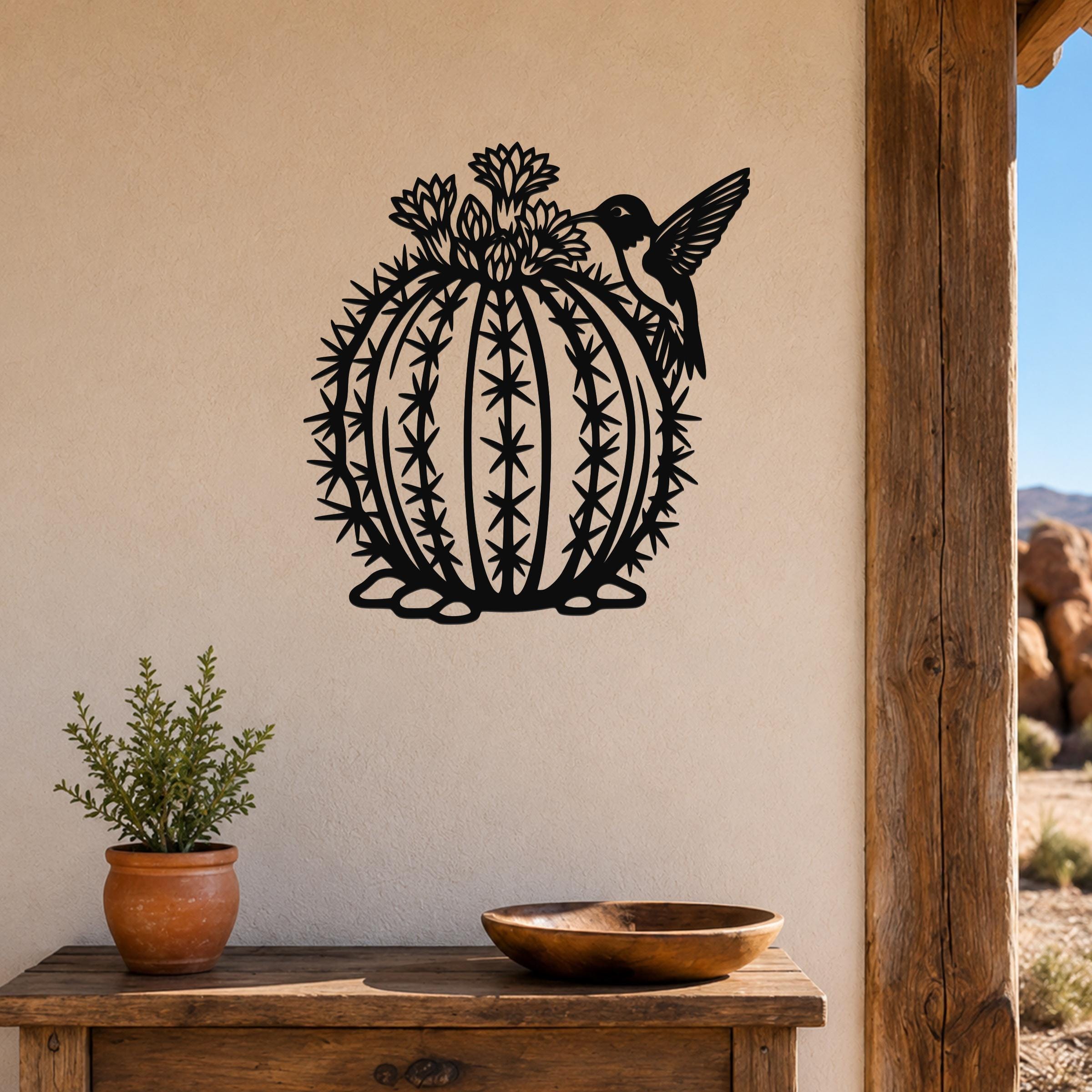 Black metal barrel cactus wall sculpture with flower and hummingbird cutouts, mounted on a beige wall above a wooden table.