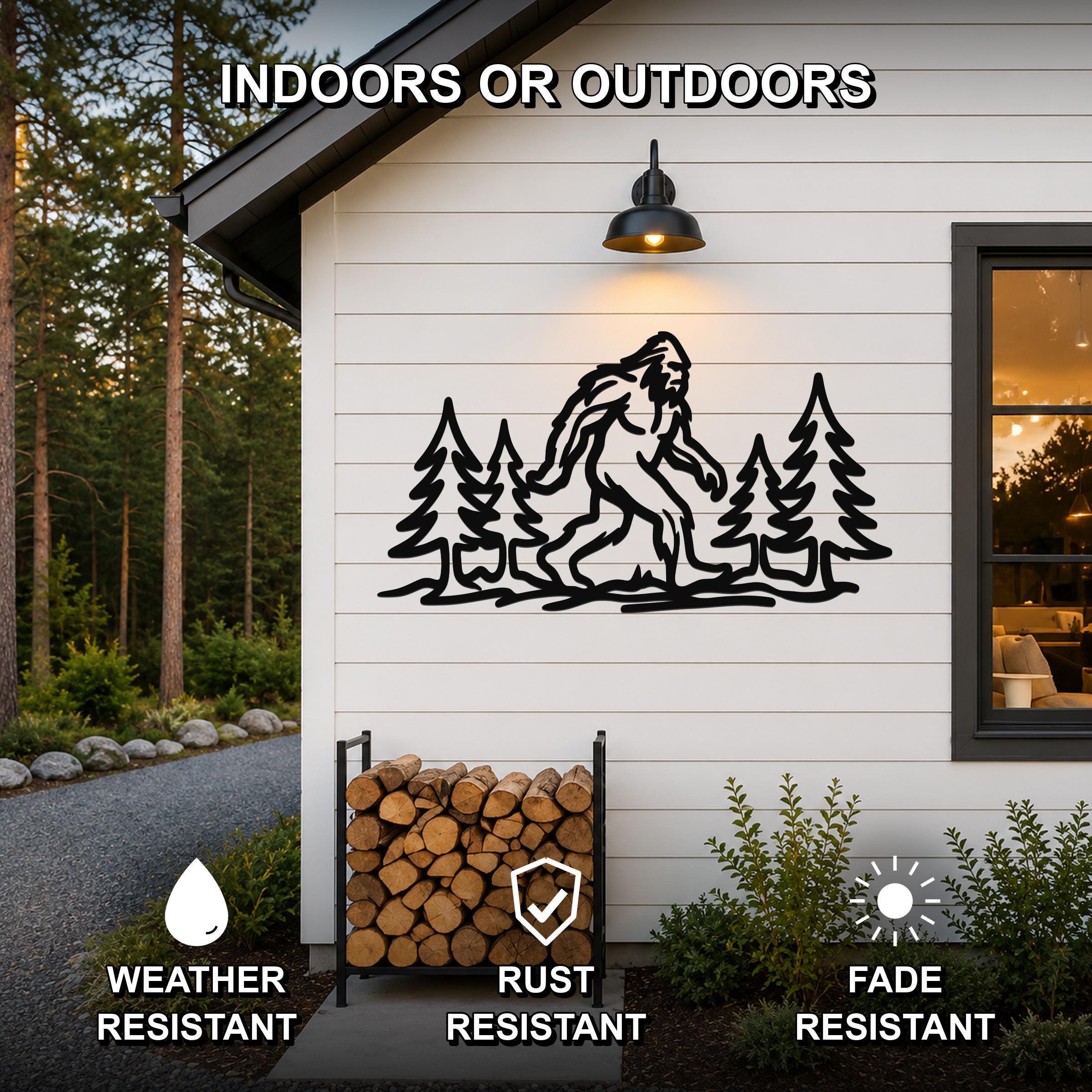 Black metal wall art featuring a Bigfoot silhouette among pine trees, mounted on a white siding exterior wall above a firewood rack, with text indicating it is weather, rust, and fade resistant for indoor or outdoor use.