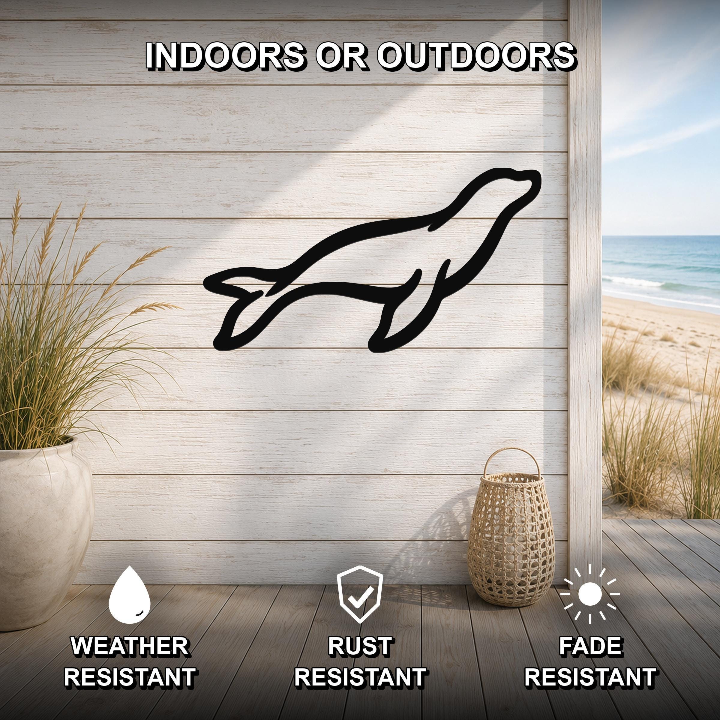 Black metal sea lion silhouette wall art mounted on a light wood plank wall, shown in an indoor-outdoor setting with a beach view, weather resistant, rust resistant, and fade resistant.