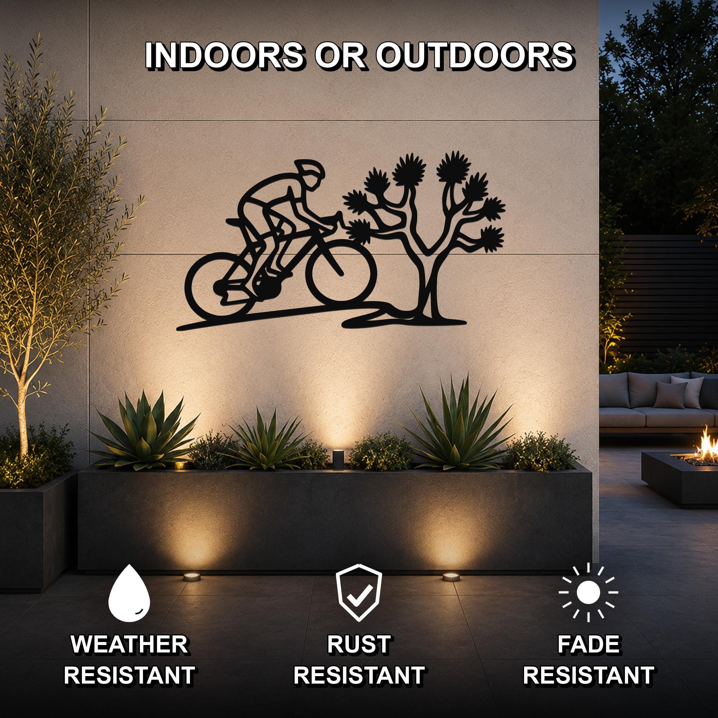 Black metal wall art featuring a cyclist silhouette and a Joshua tree cutout, mounted on an outdoor wall above a planter in a modern patio setting.