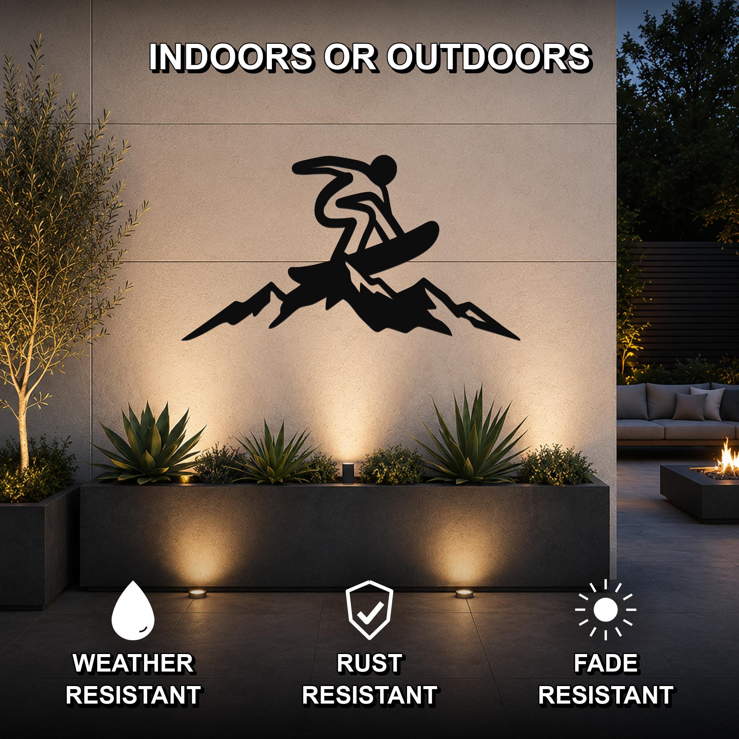 Black metal wall art featuring a snowboarder silhouette above mountain peaks, mounted on an outdoor wall above a planter in a modern patio setting.