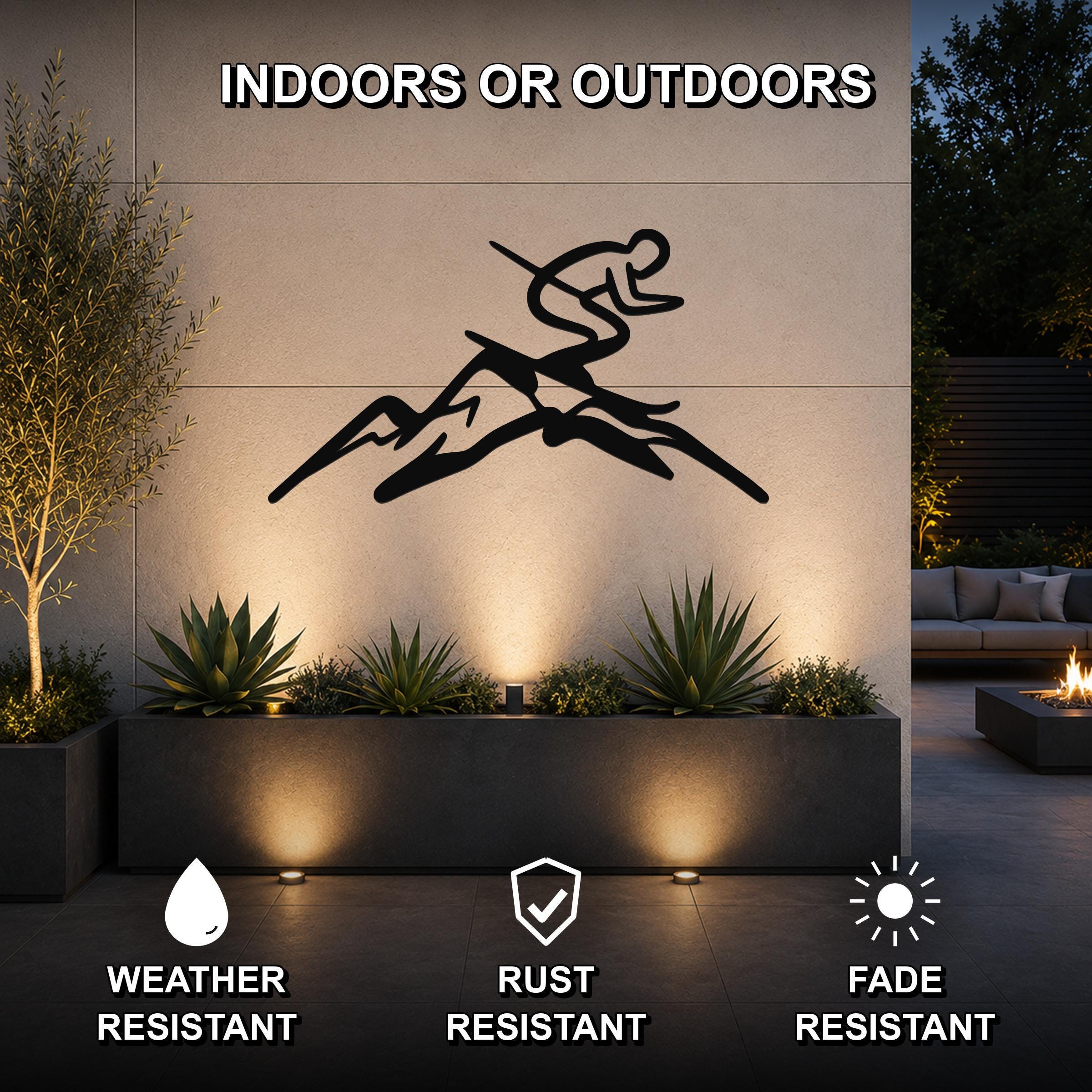 Black metal wall art featuring a skier silhouette in motion above mountain peaks, mounted on an outdoor wall above a planter with warm uplighting, weather resistant, rust resistant, and fade resistant.