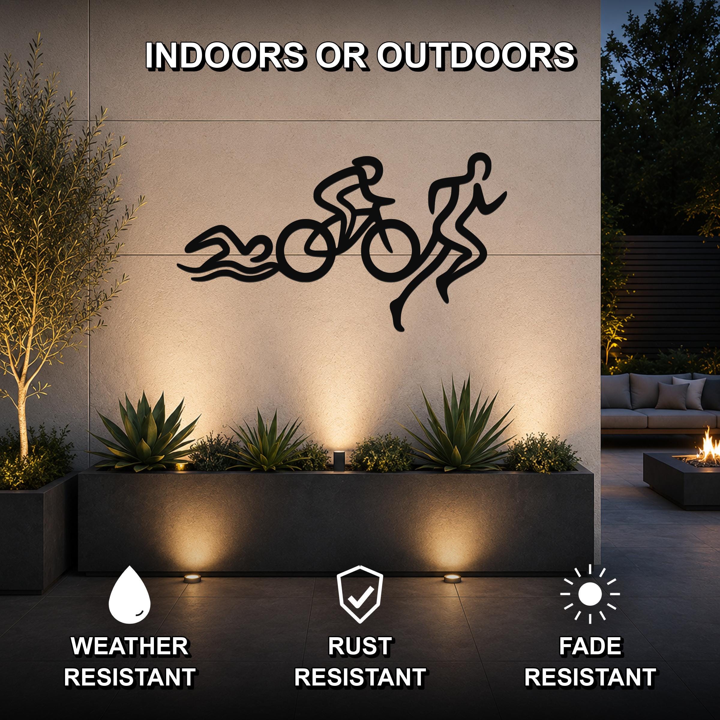 Black metal triathlon wall art with swim, bike, and run silhouette cutouts, mounted on an outdoor patio wall above a planter with accent lighting.