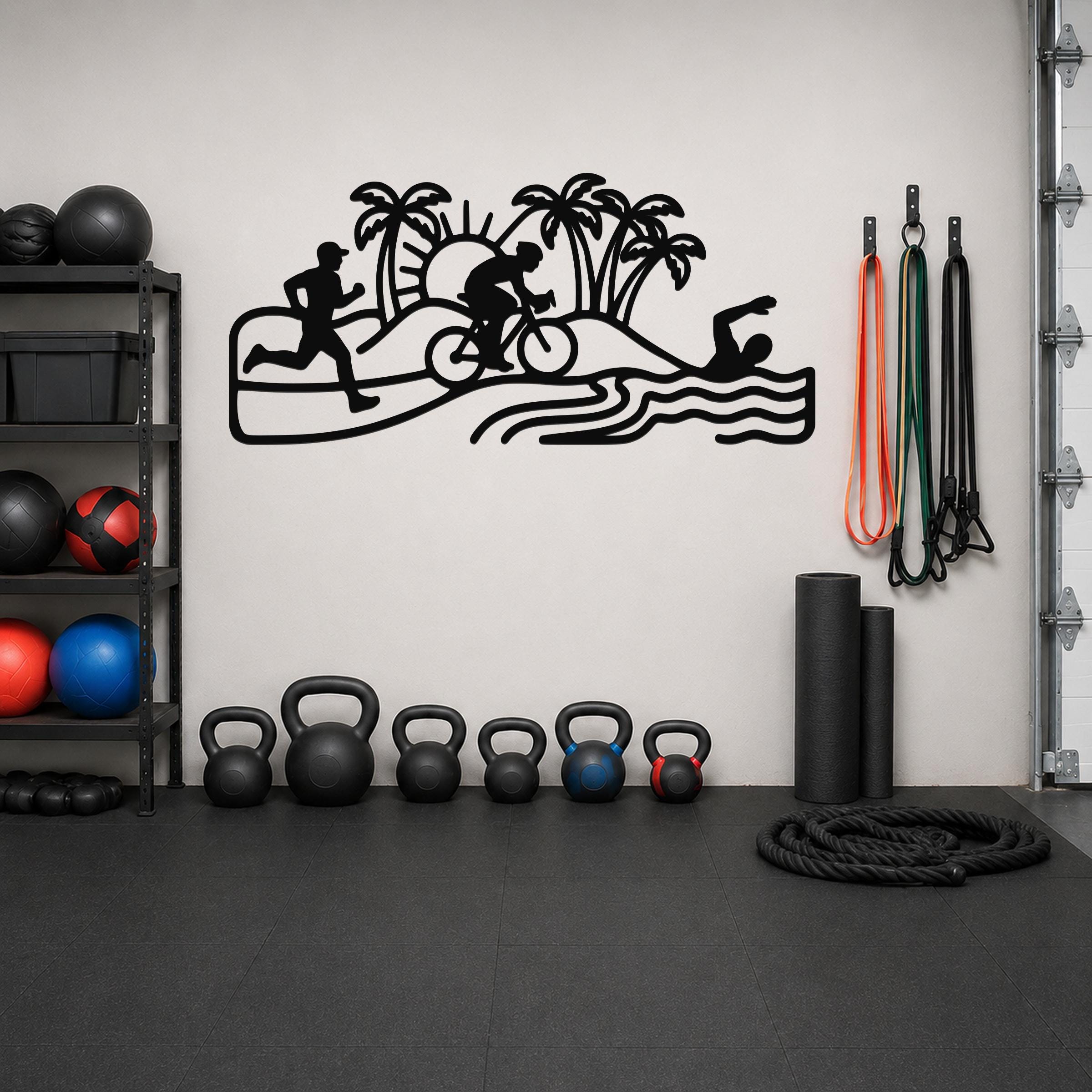 Black metal wall art cutout of a triathlon scene with swim, bike, and run silhouettes, palm trees, and waves, mounted on a gym wall above a row of kettlebells.