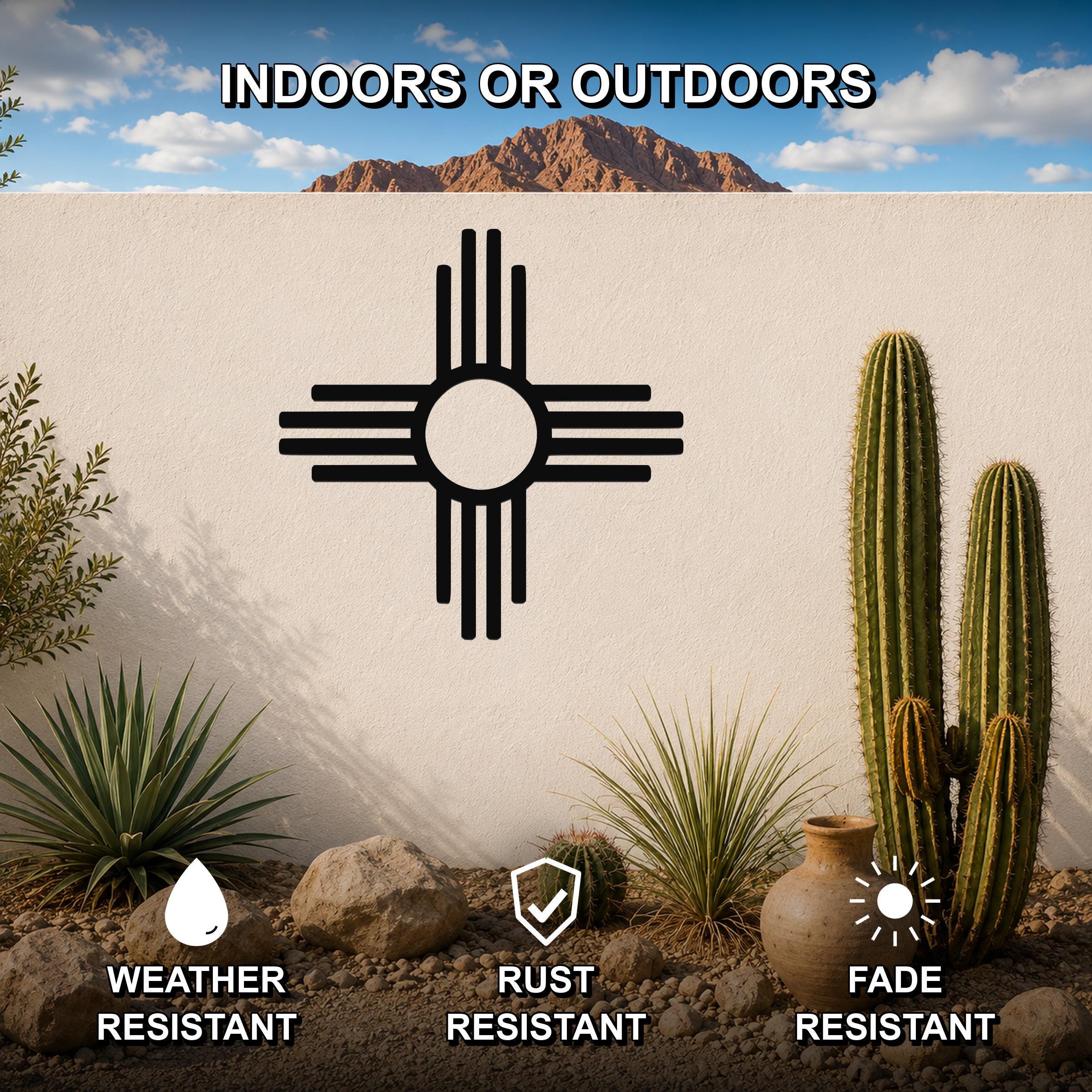 Black metal Zia Sun symbol wall art with a bold circular design and radiating lines, mounted on a white outdoor wall in a desert garden setting with cacti and rocks.