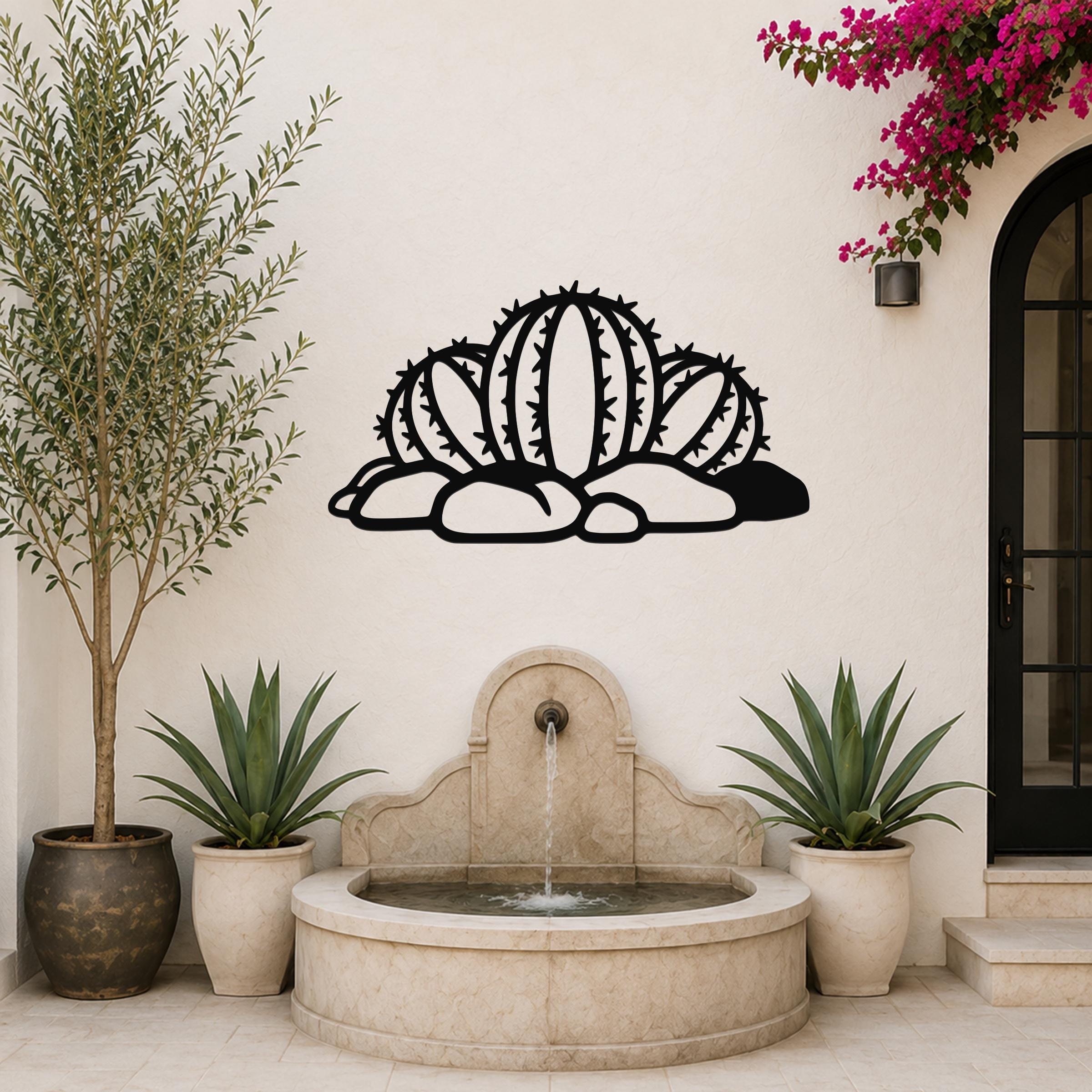 Black metal barrel cactus wall art with spines and rocks cutout, mounted on a white outdoor wall above a stone fountain.