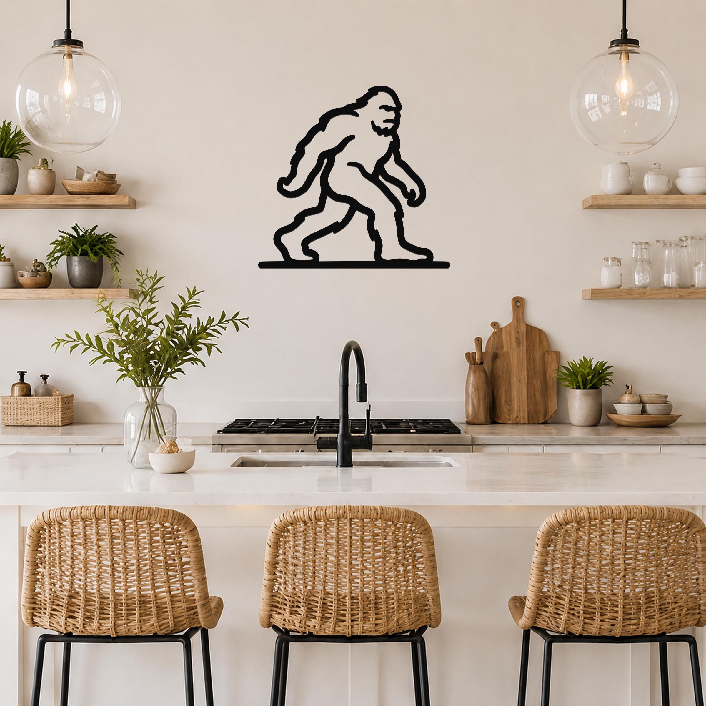 Black metal Bigfoot wall art silhouette mounted on a kitchen wall above a counter, with wicker bar stools and open shelves visible below.