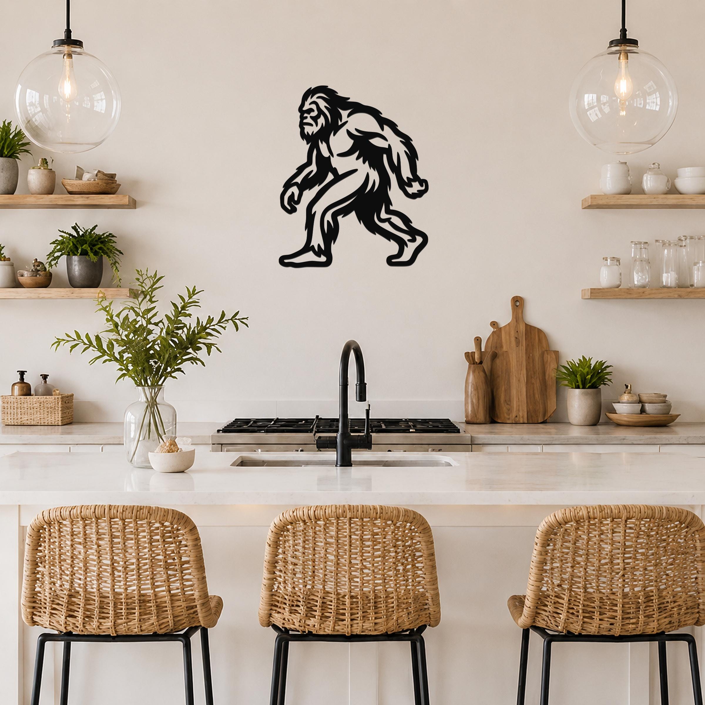Black metal Bigfoot silhouette wall art mounted on a kitchen wall above a counter, with wicker bar stools and open shelving visible.