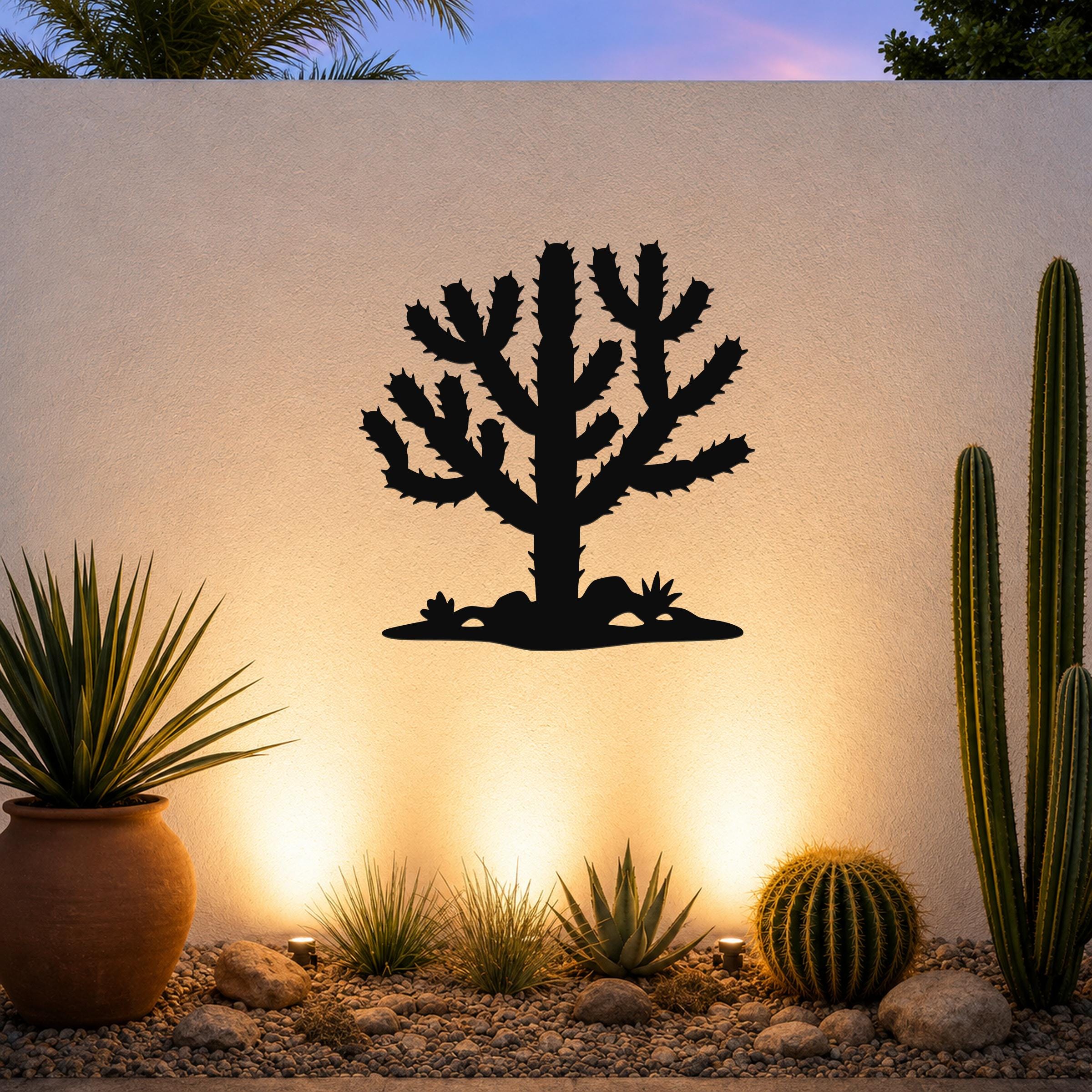 Black metal Cholla cactus wall art silhouette with spiny branches and ground cutout, mounted on a textured outdoor wall above a desert garden with potted plants and ground lights.
