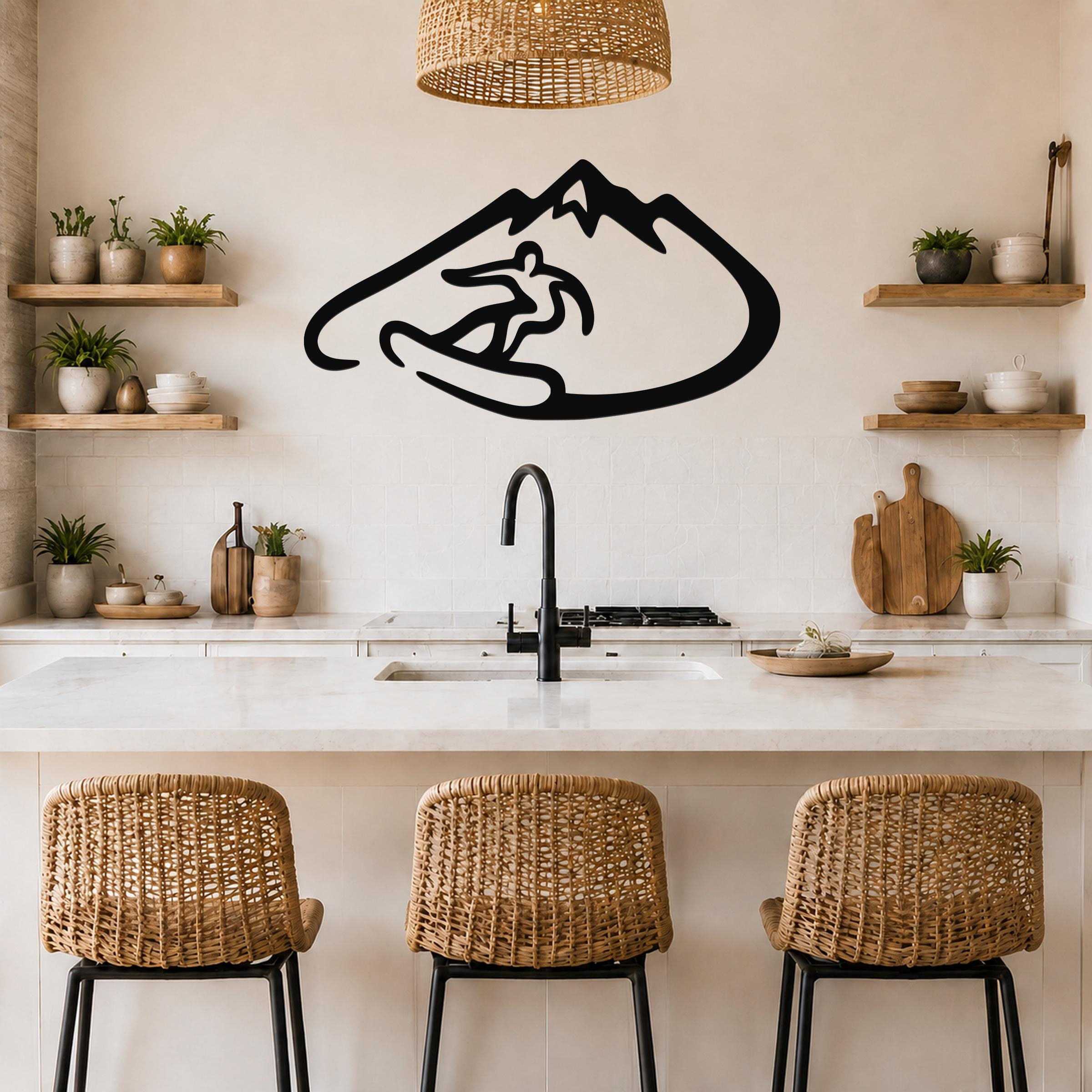 Black metal wall art featuring a minimalist silhouette of a snowboarder in a mountain, mounted above a kitchen counter with wicker stools and wooden shelves.