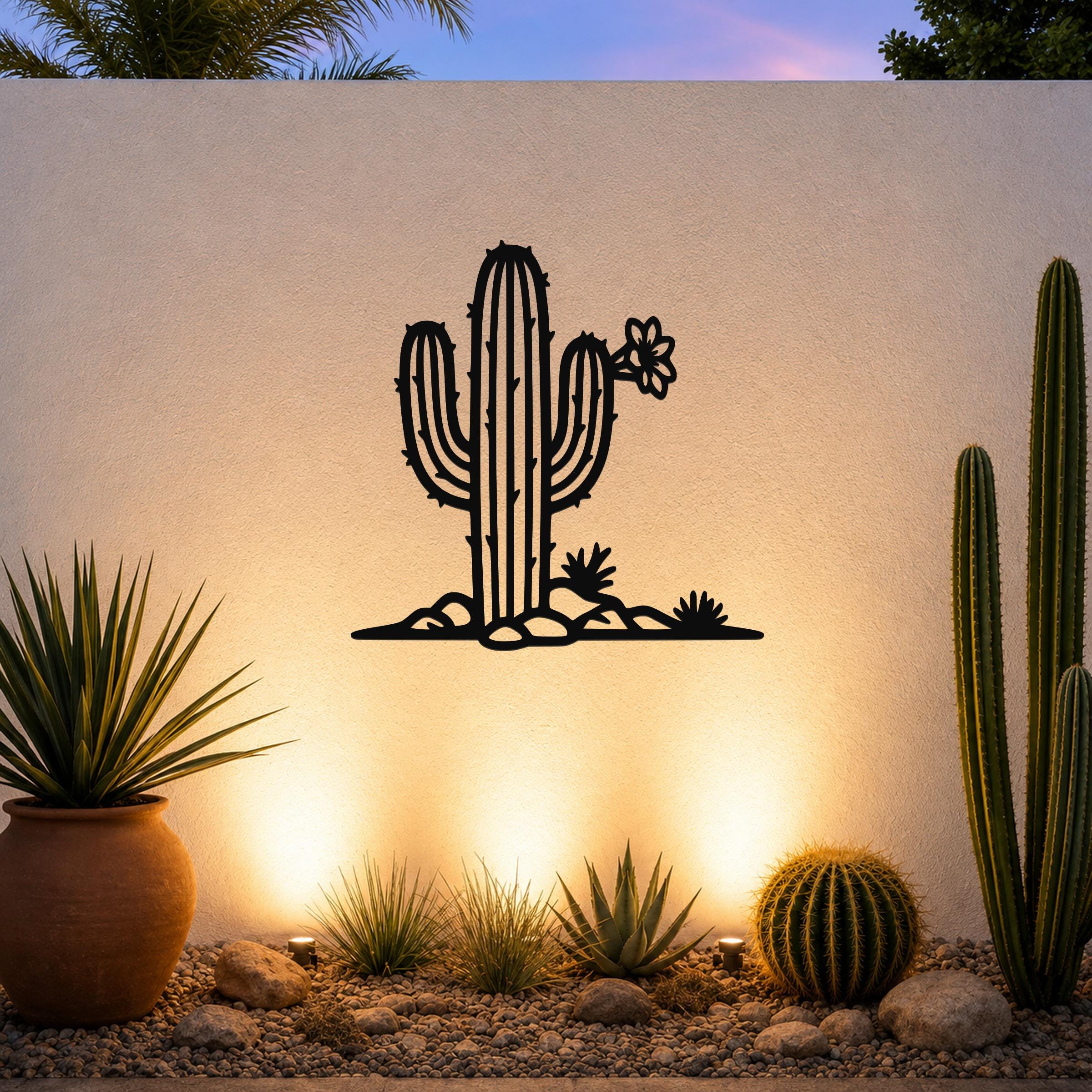Black metal wall art silhouette of a saguaro cactus with flower and smaller cacti, mounted on an outdoor wall above a gravel garden with potted plants and uplighting.