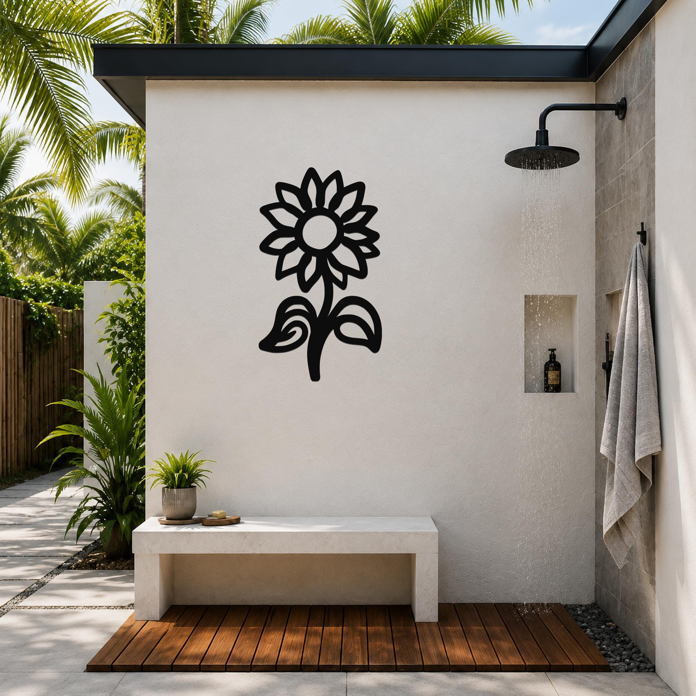Black metal sunflower wall art with detailed petal and leaf cutouts, mounted on a white outdoor shower wall above a concrete bench.