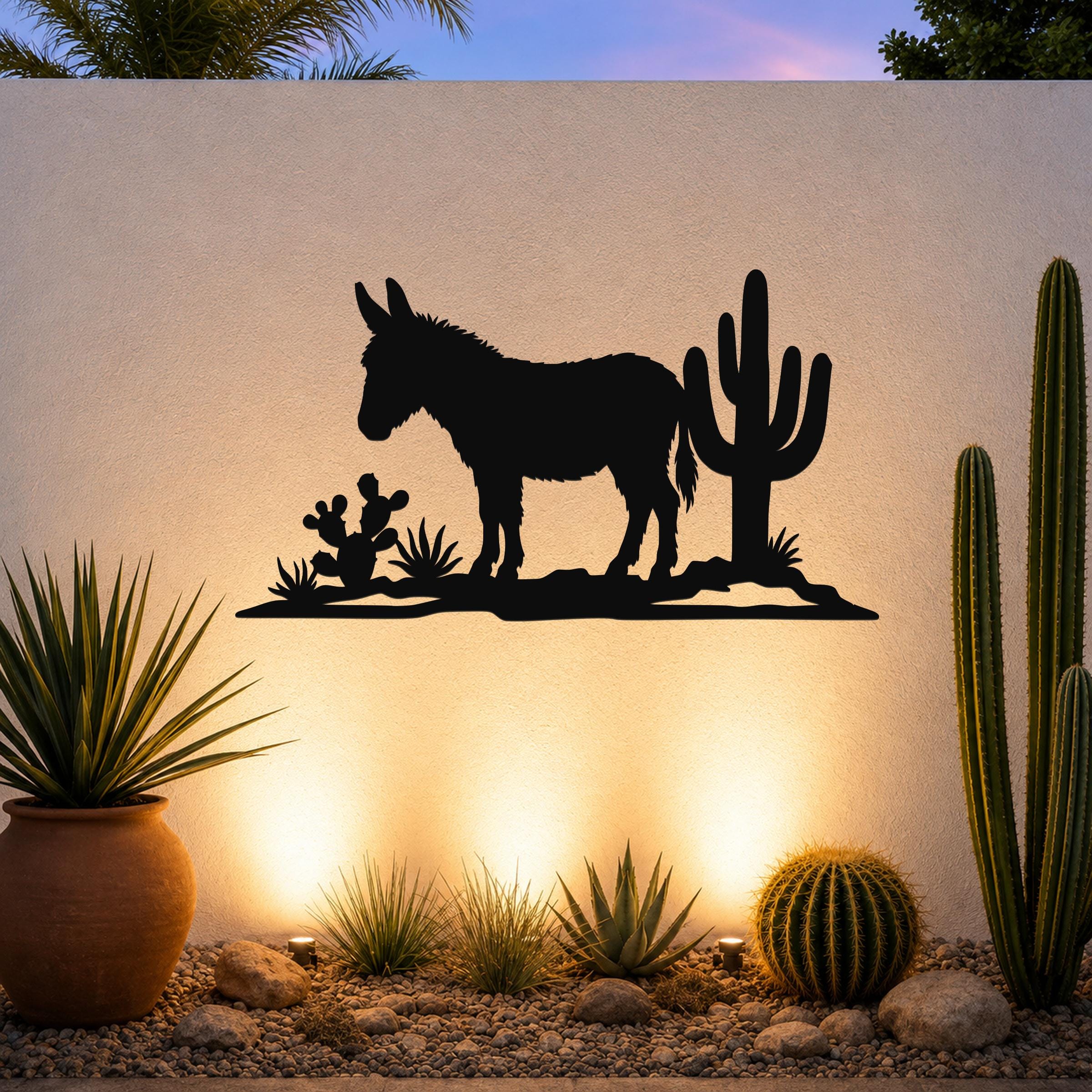 Black metal wall art silhouette of a burro and cacti, mounted on an outdoor wall above a desert garden with warm uplighting.