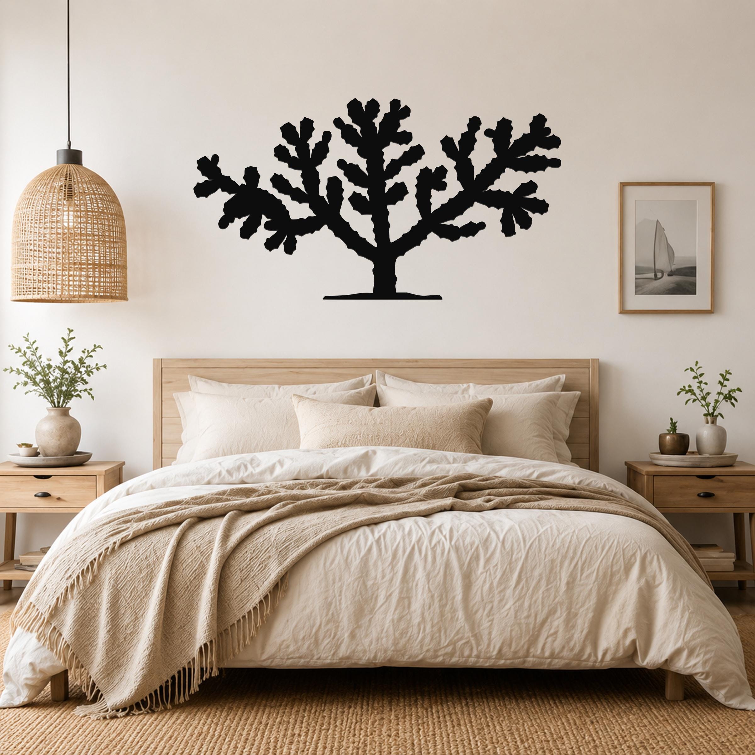 Black metal Cholla cactus wall art silhouette with branching arms, mounted above a wooden bed in a neutral bedroom.