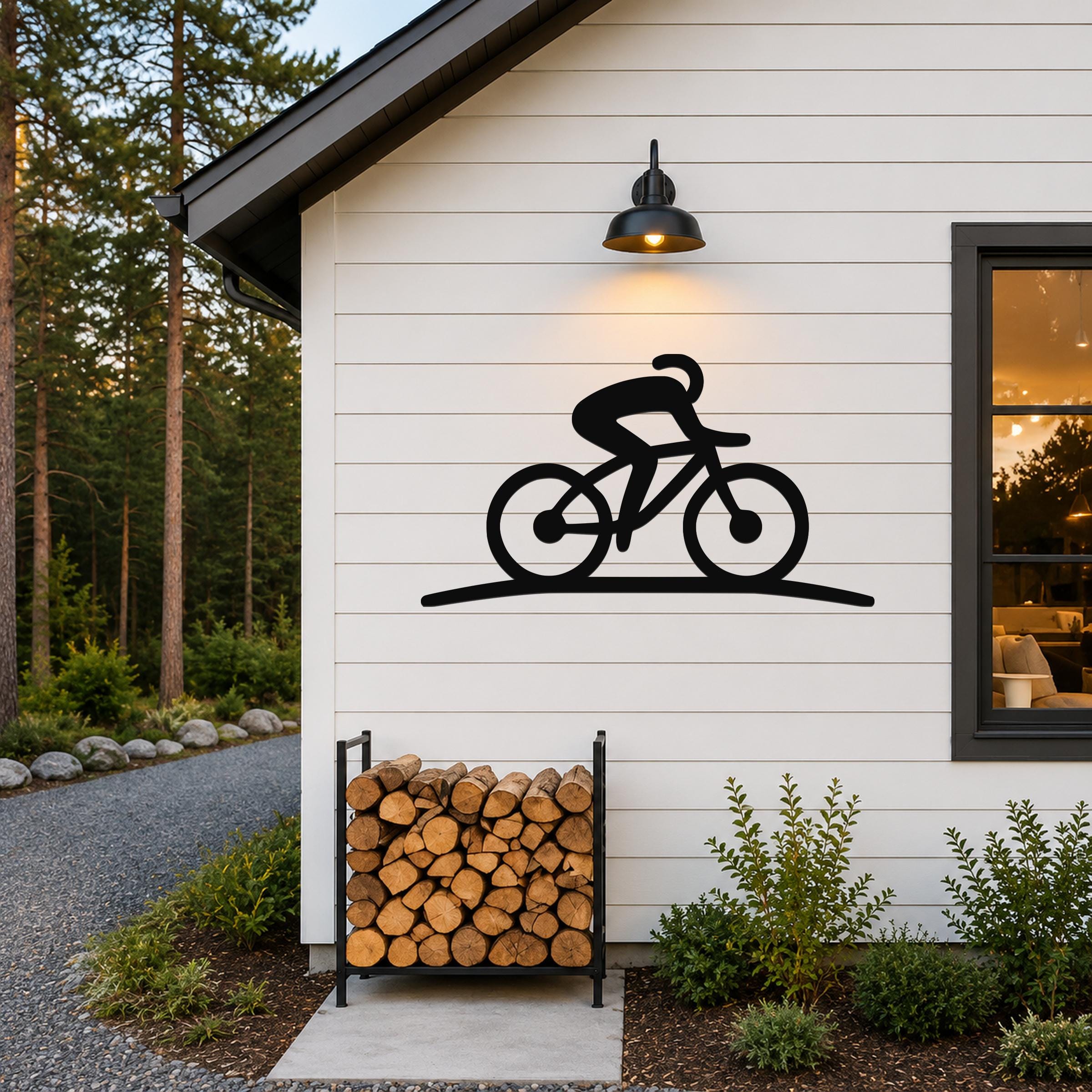 Black metal cycling wall art silhouette mounted on a white siding wall above a firewood rack, illuminated by a wall sconce.