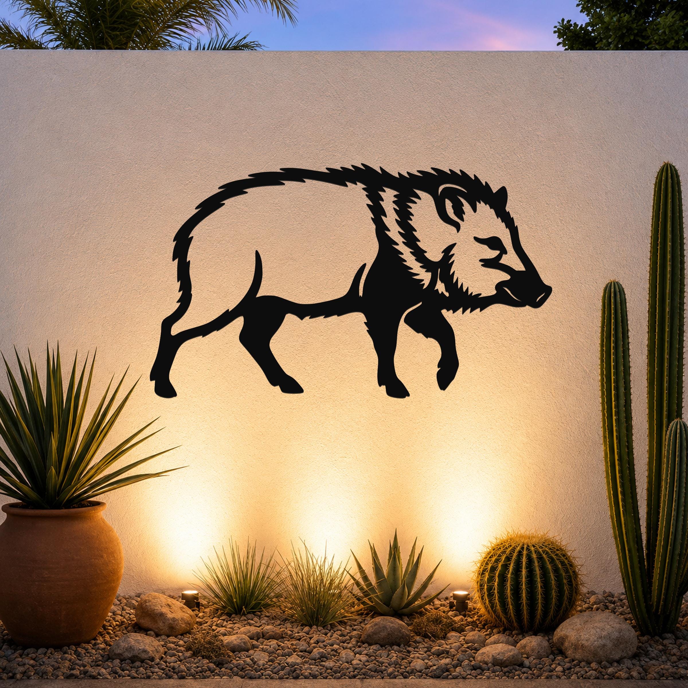 Black metal javelina wall art silhouette mounted on a textured outdoor wall above a desert garden with cacti and warm uplighting.