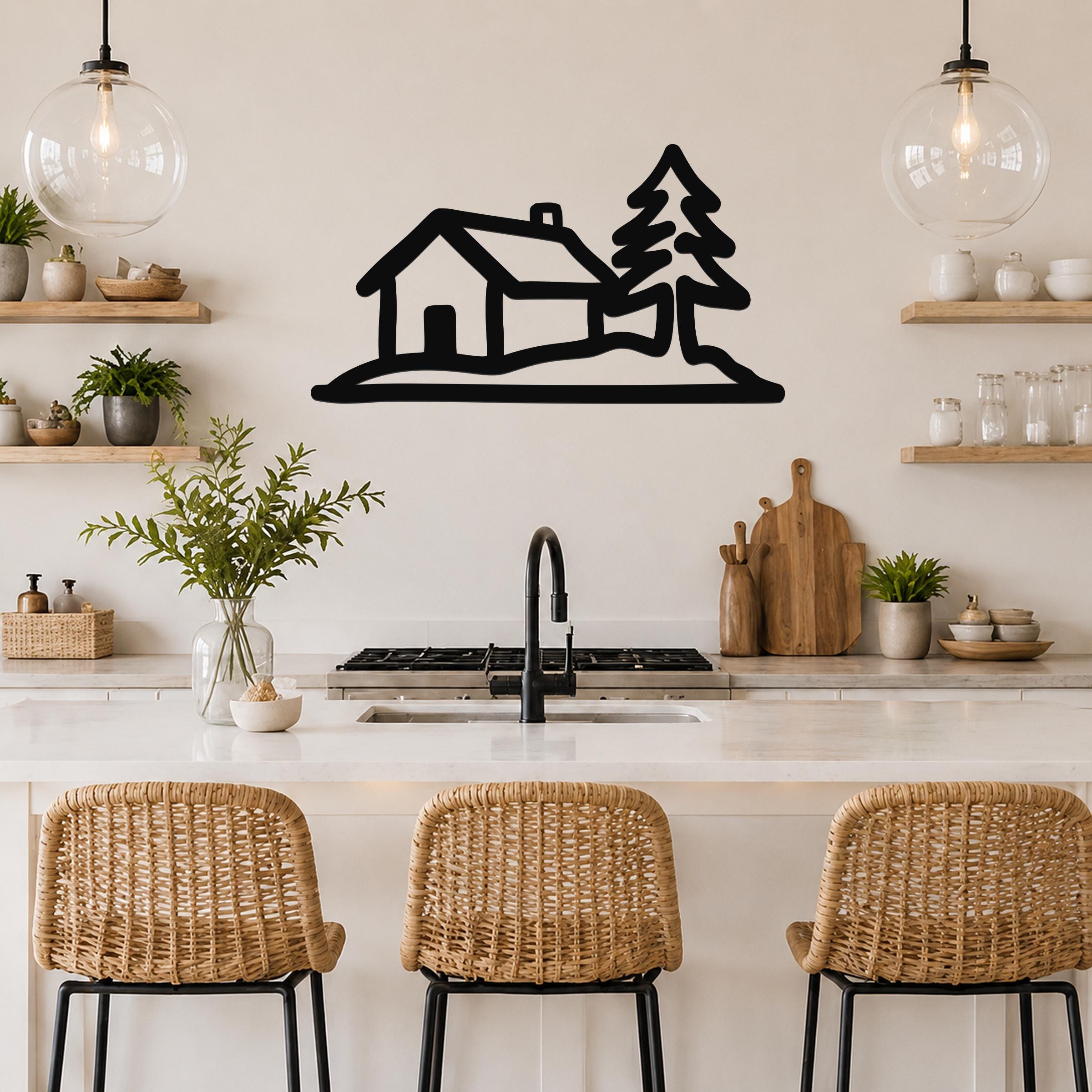 Black metal wall art silhouette of a cabin and pine tree, mounted above a kitchen counter with wicker stools and open shelving.