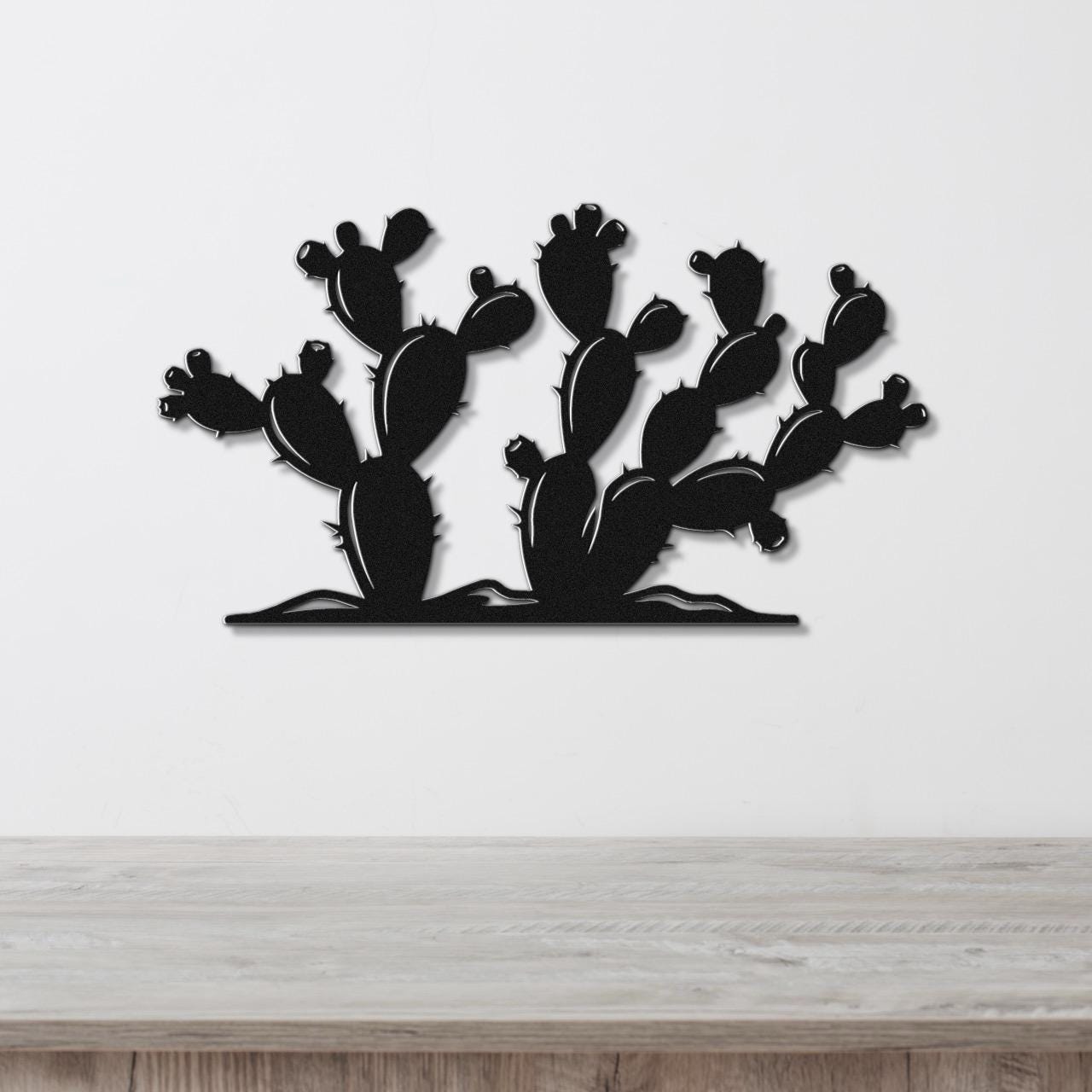 Black metal wall art cutout of a prickly pear cactus with multiple pads and spines, mounted on a white wall above a light wood surface.