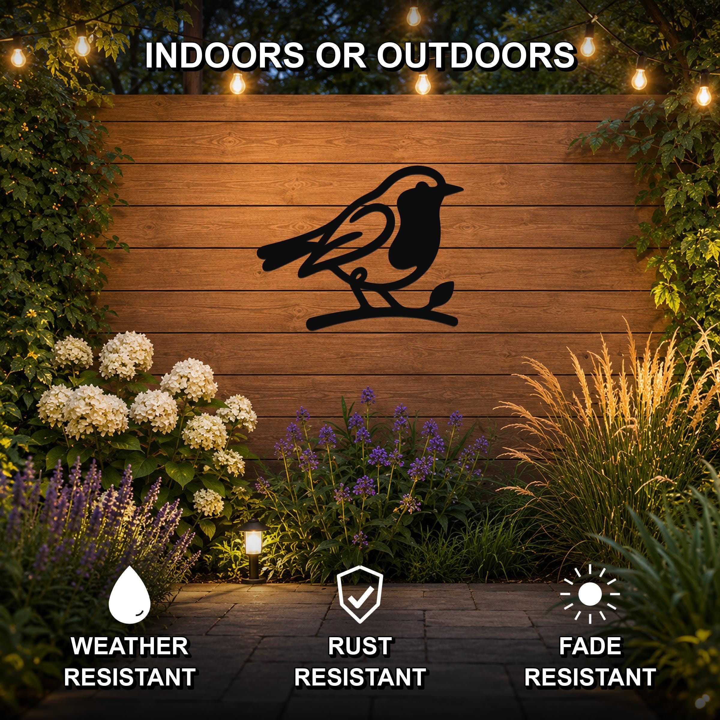 Black metal robin wall art silhouette perched on a branch, mounted on a wooden outdoor wall above a garden with flowers and string lights, weather resistant, rust resistant, fade resistant.