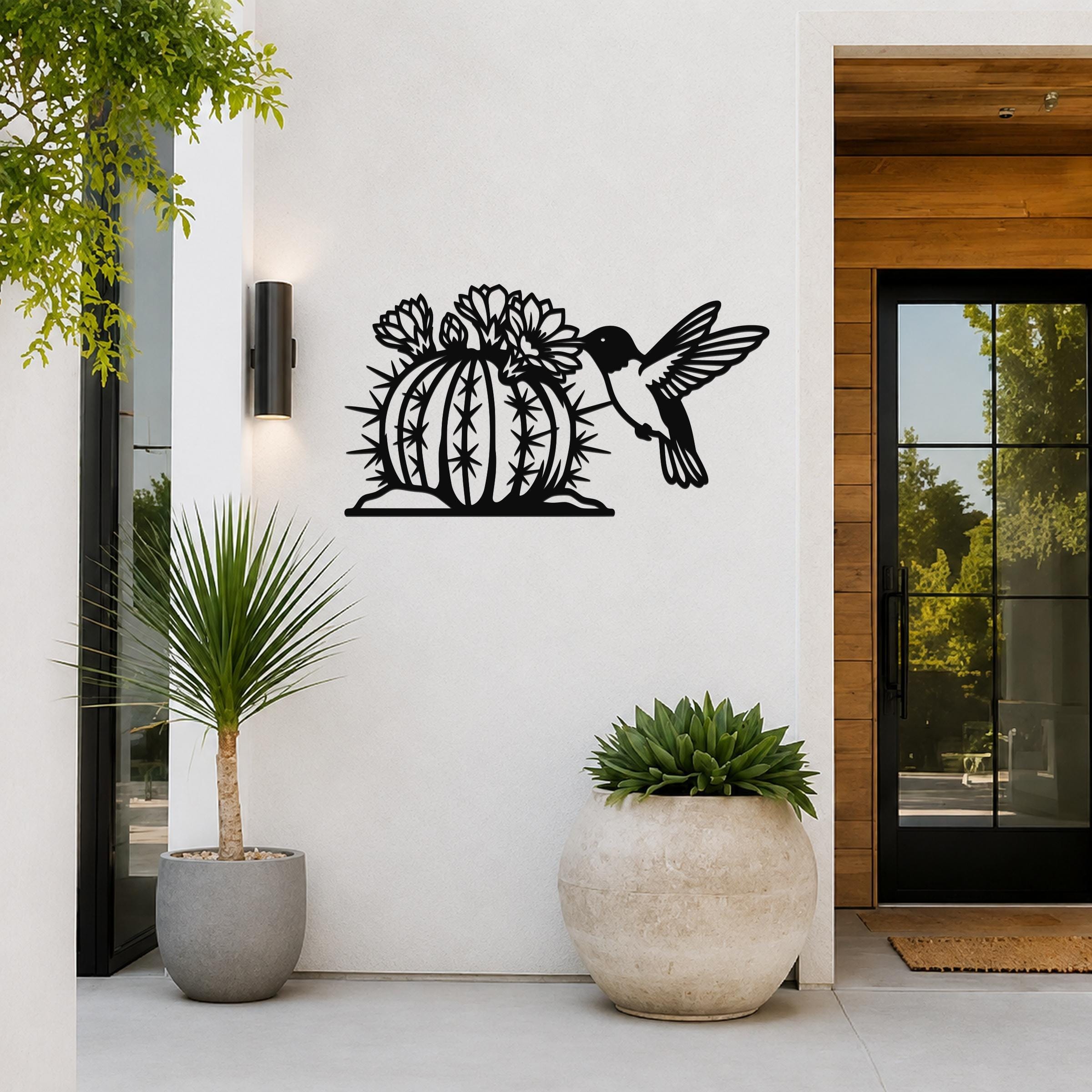 Black metal barrel cactus wall art with flowering blooms and a hummingbird cutout, mounted on a white exterior wall beside a glass door and potted plants.