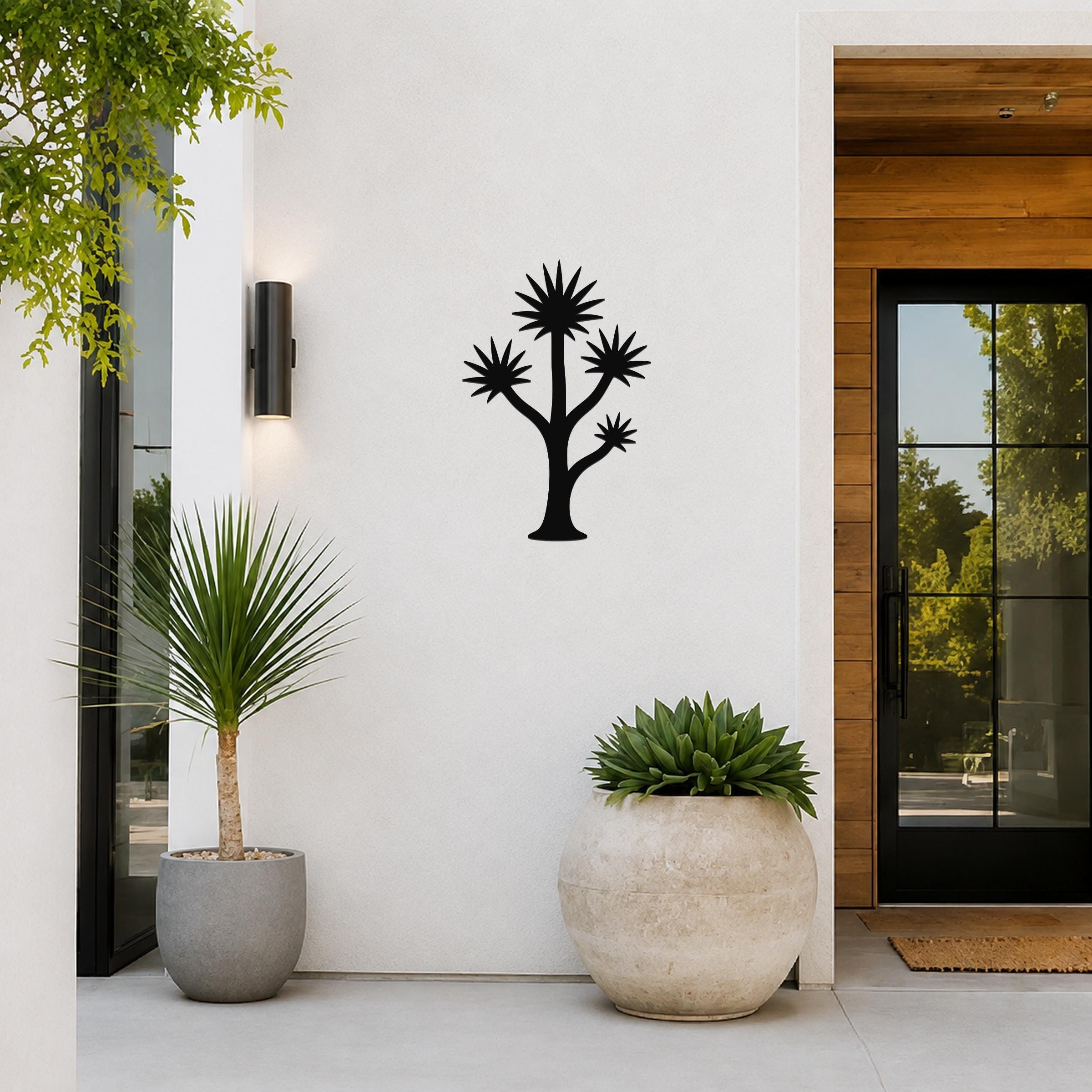 Black metal wall art silhouette of a yucca tree with spiky leaf clusters, mounted on a white exterior wall beside a glass door and potted plants.