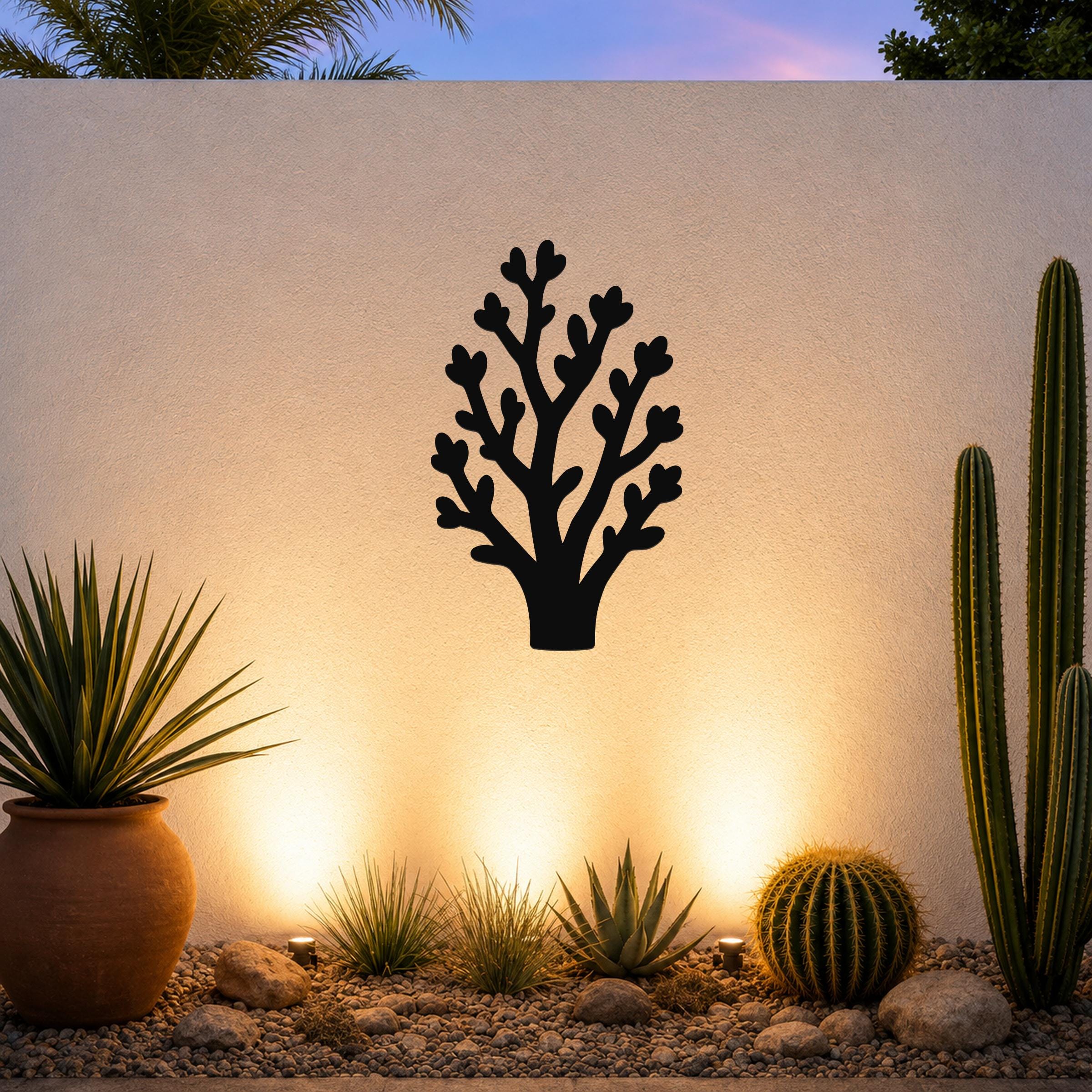 Black metal wall art in a tree silhouette with branching cutouts, mounted on an outdoor wall above a desert garden with cacti and warm uplighting.