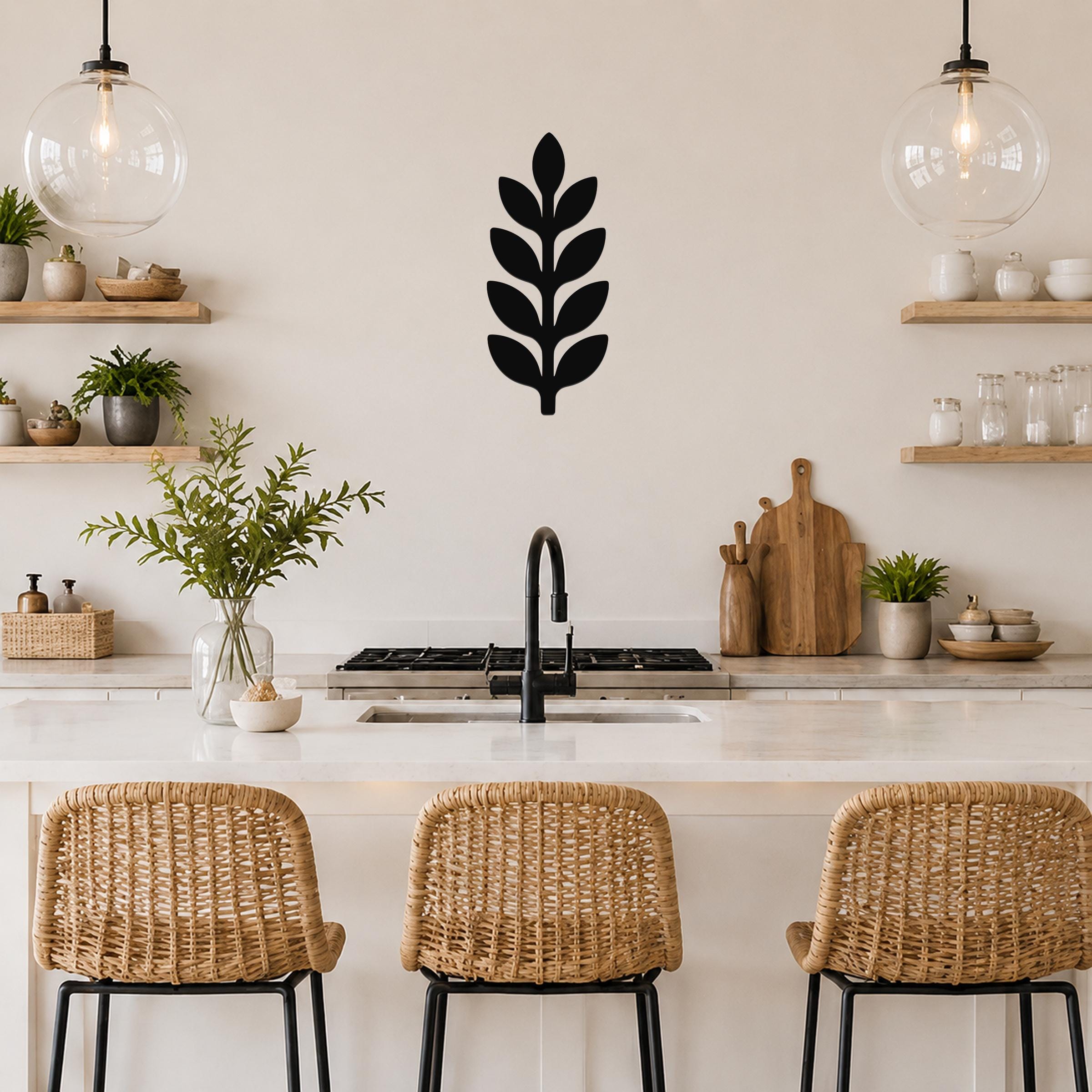 Black metal wall art in a leafy sprig silhouette, mounted on a kitchen wall above a counter with pendant lights and woven bar stools.