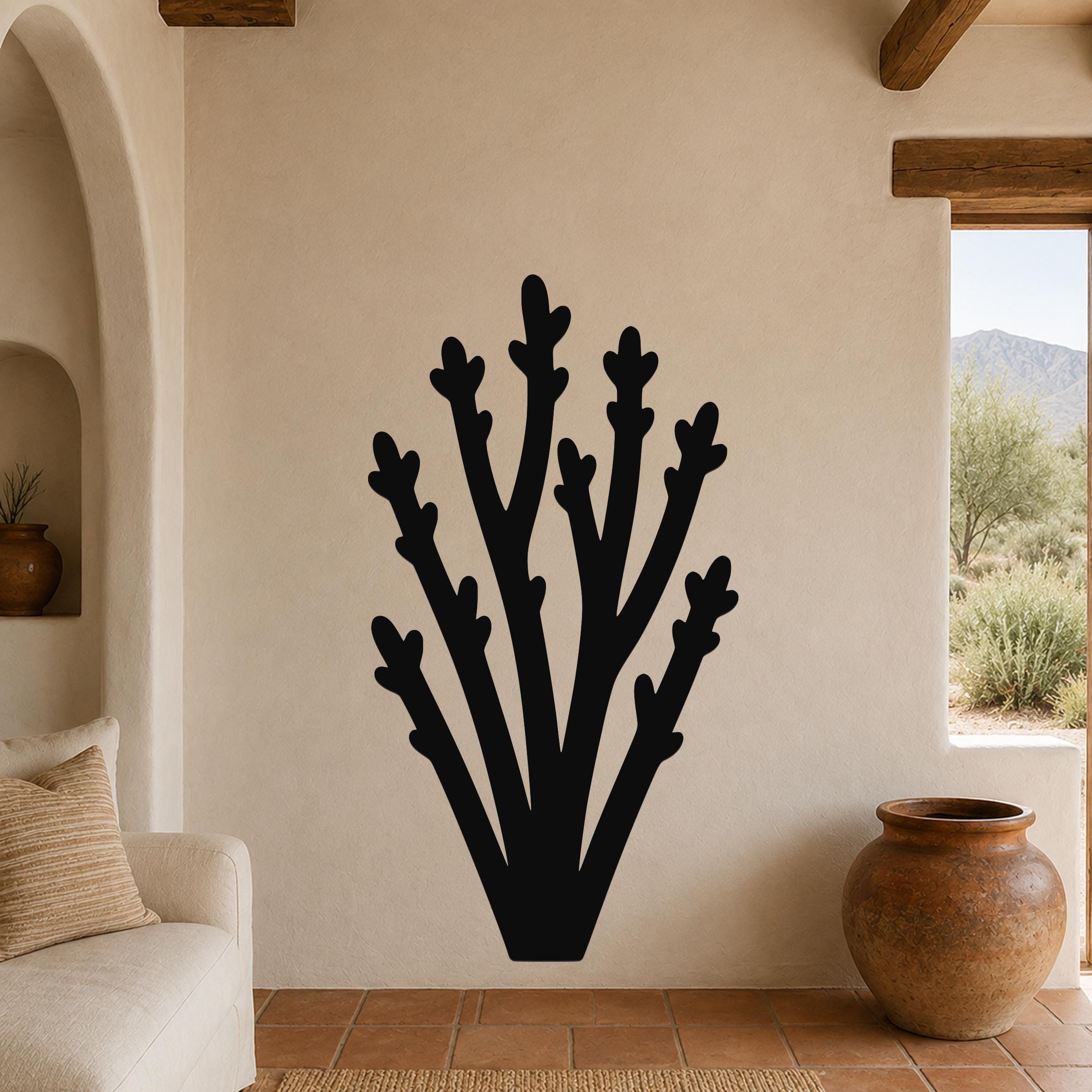Black metal wall art in a desert branch silhouette, mounted on a beige stucco wall in a living room with terracotta flooring and wooden beams.