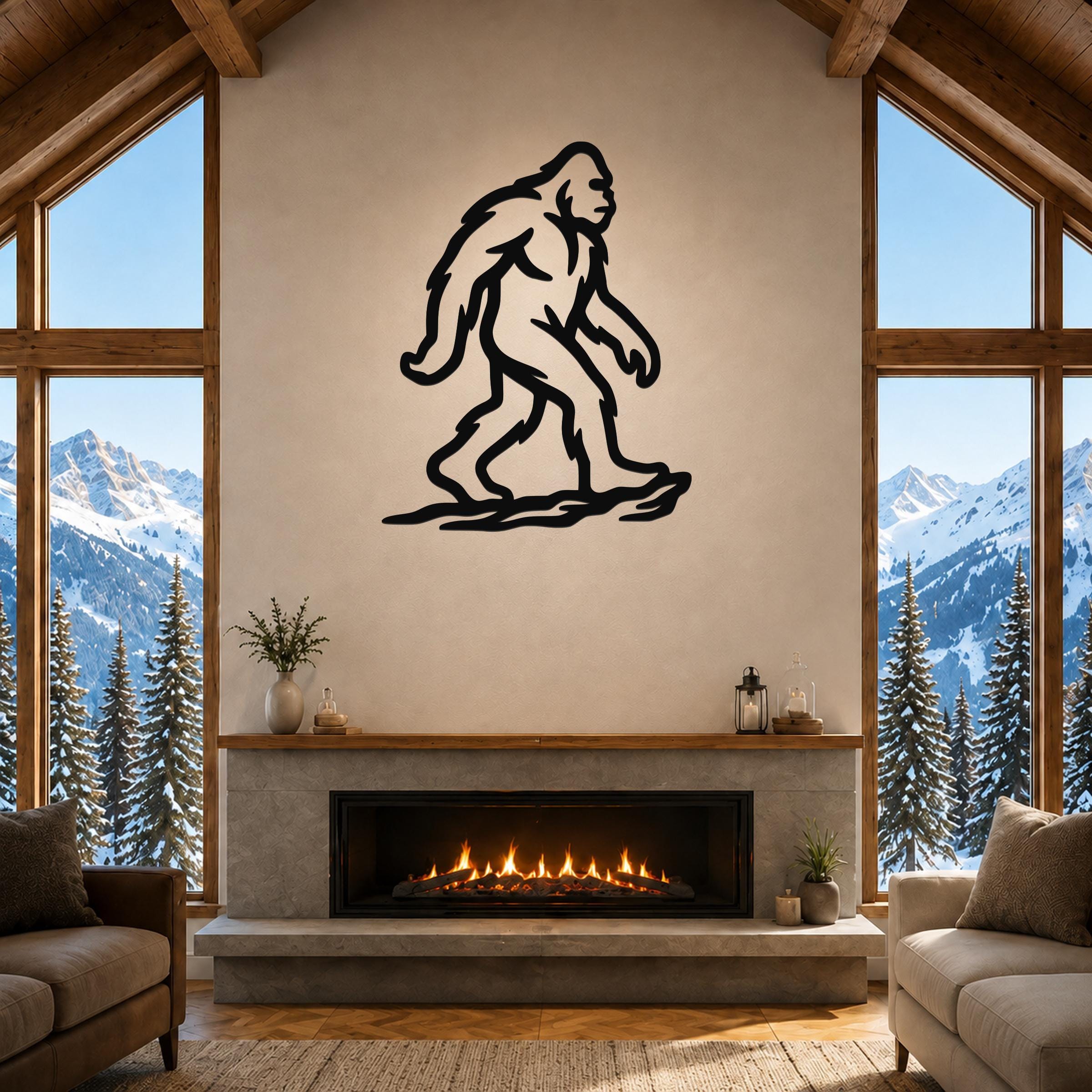Black metal Bigfoot silhouette wall art mounted above a fireplace in a rustic living room with large windows and snowy mountain views.