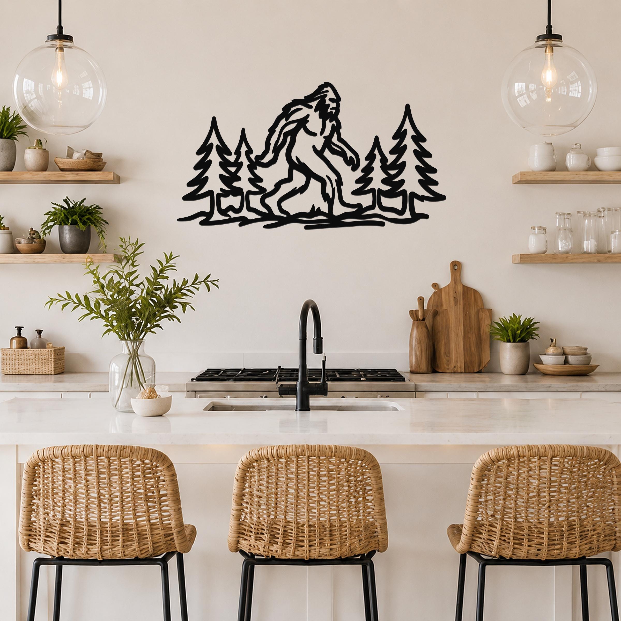 Black metal wall art featuring a Bigfoot silhouette among pine trees, mounted above a kitchen counter with wicker bar stools.