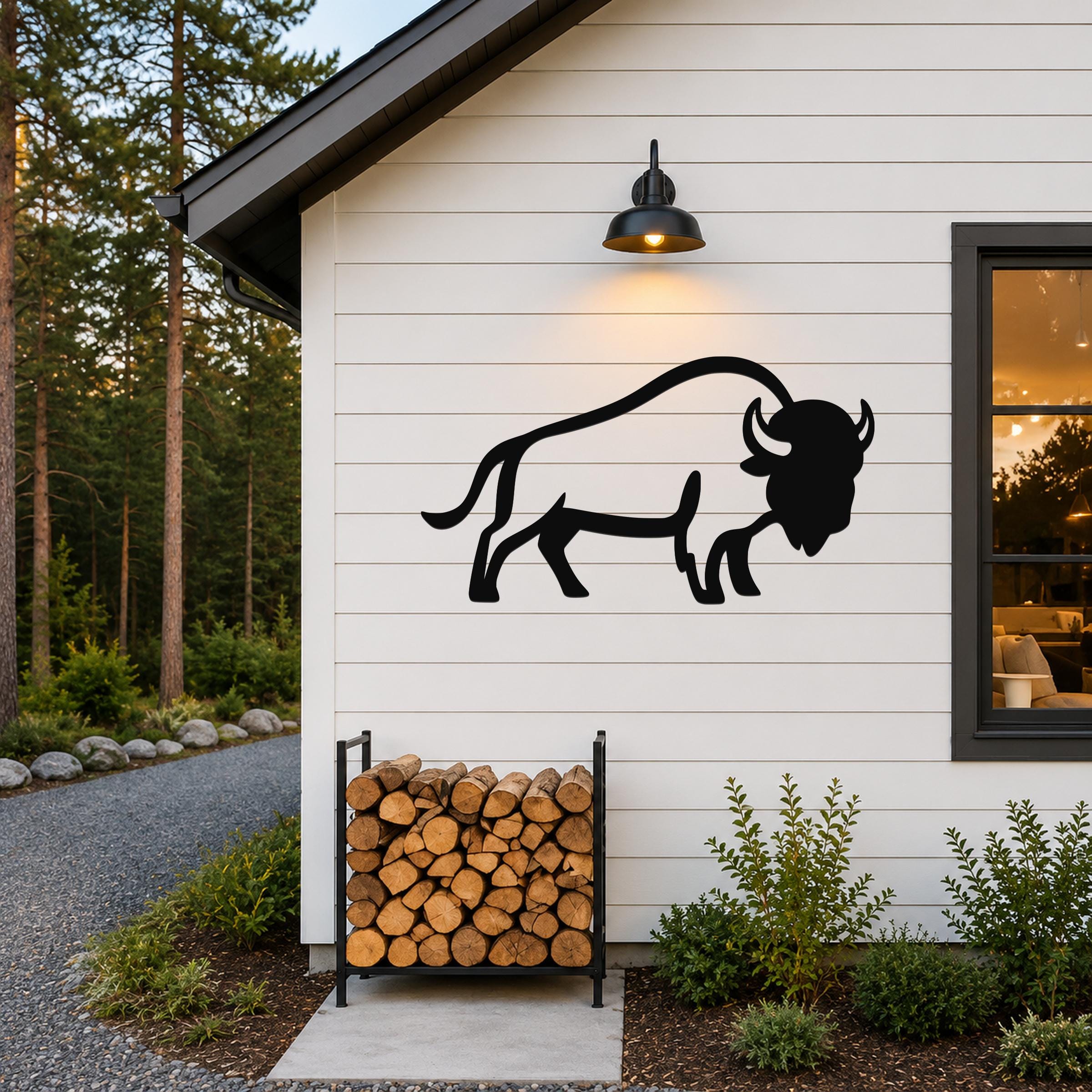 Black metal buffalo silhouette wall art mounted on a white horizontal siding wall above a firewood rack, with a lit outdoor sconce above and a window to the right.