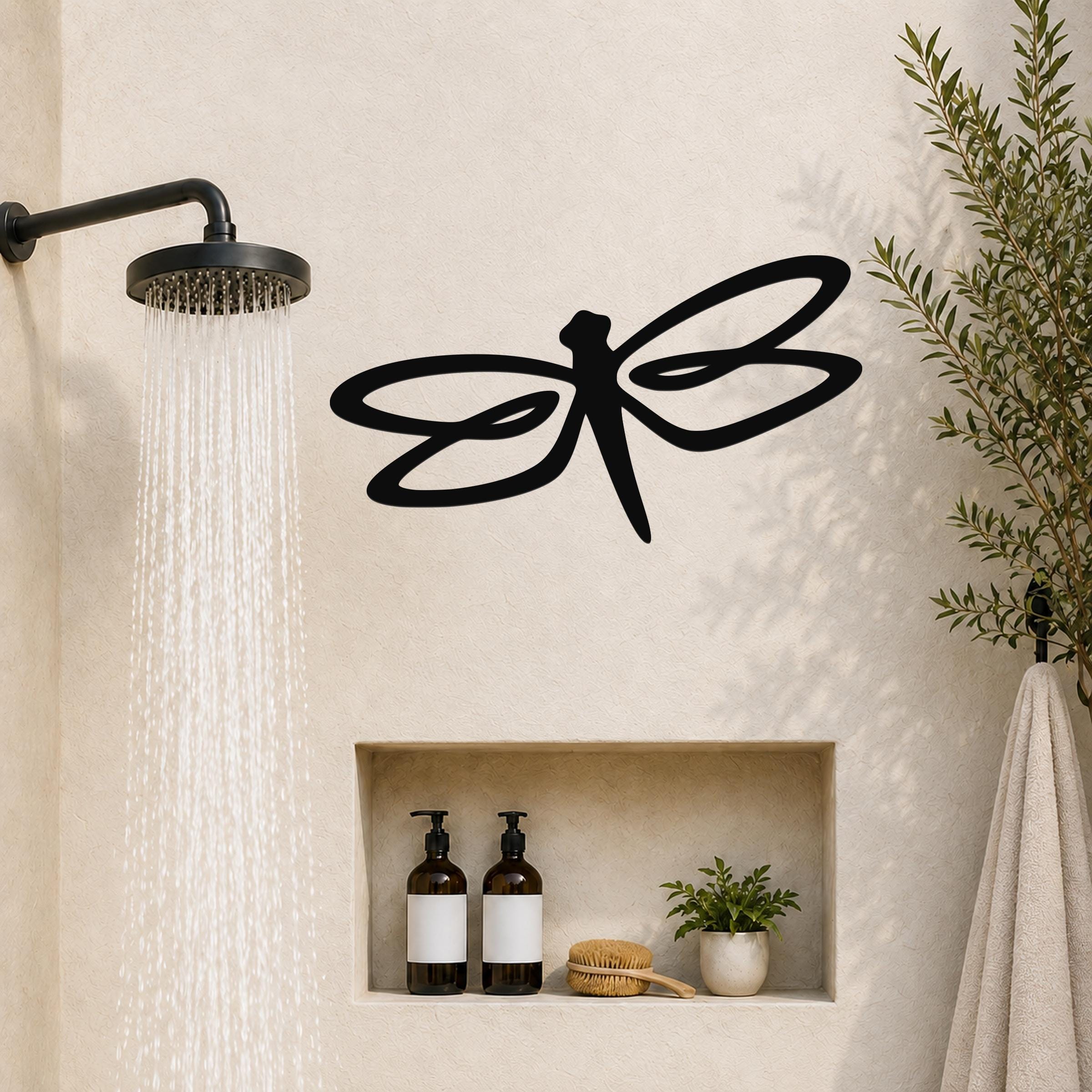 Black metal dragonfly wall art with flowing wing cutouts, mounted on a bathroom wall above a built-in shelf.