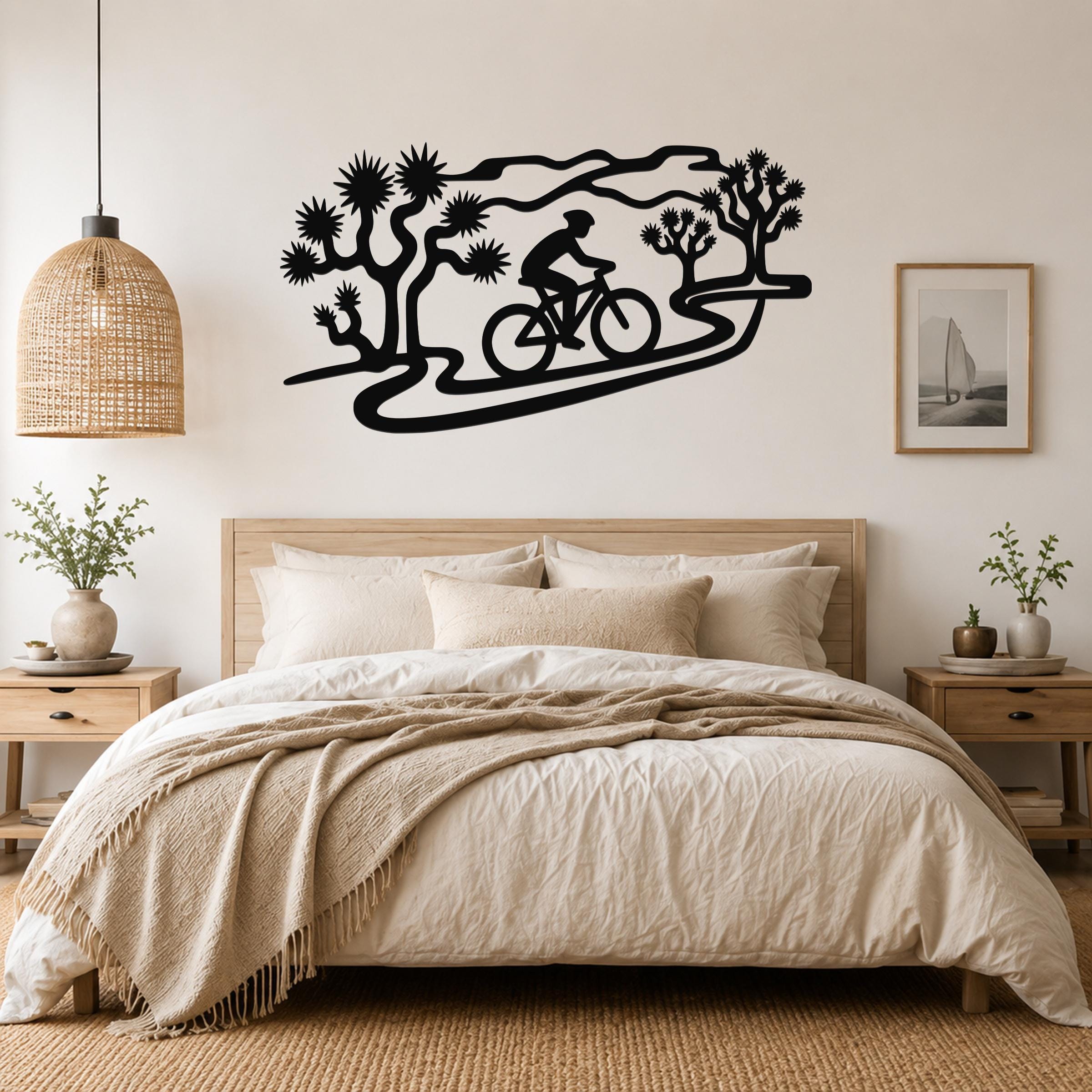 Black metal wall art silhouette of a cyclist on a winding path through Joshua trees, mounted above a bed in a bedroom.