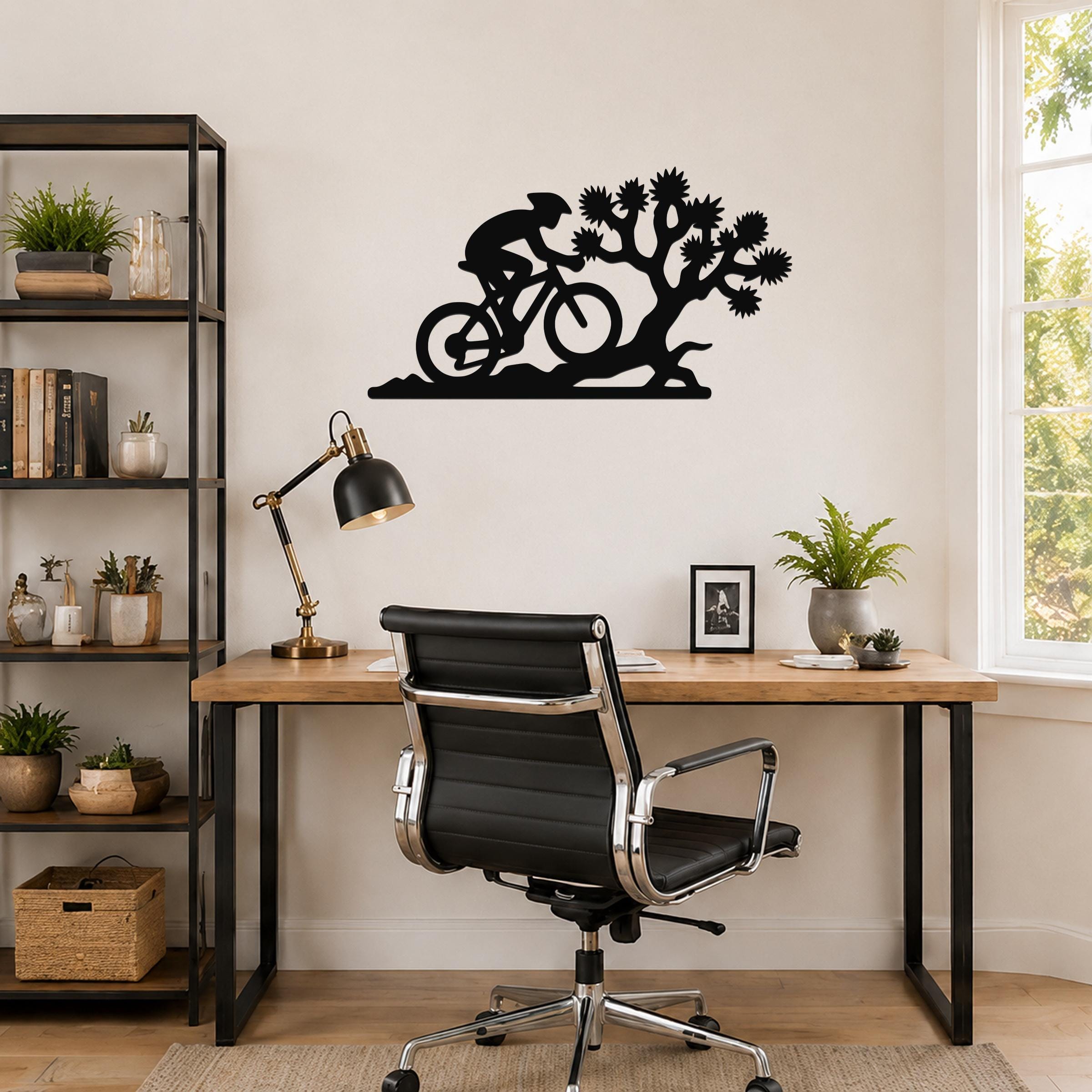 Black metal wall art silhouette of a cyclist riding past a Joshua tree, mounted above a wooden desk in a home office.