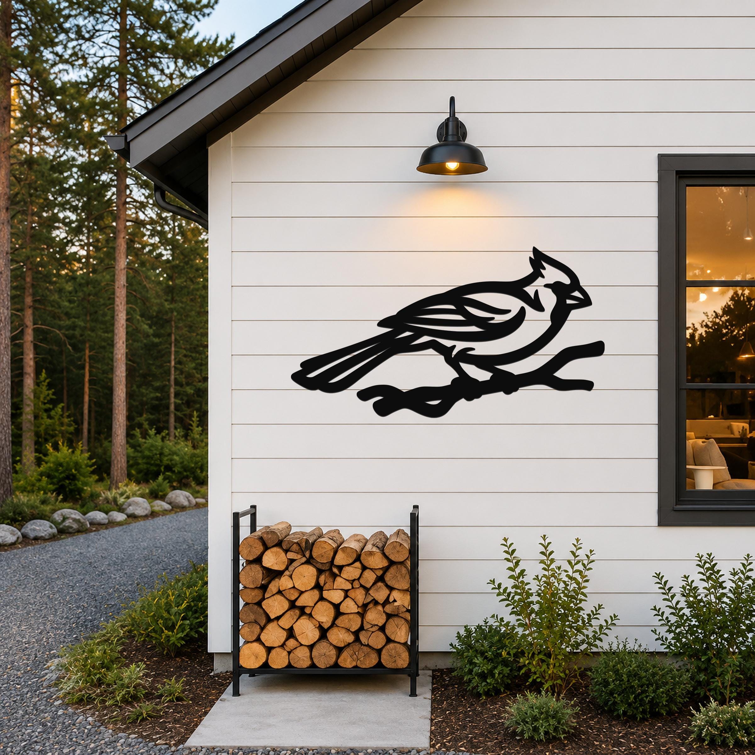 Black metal cardinal wall art in silhouette form, perched on a branch, mounted on a white exterior wall above a firewood rack.