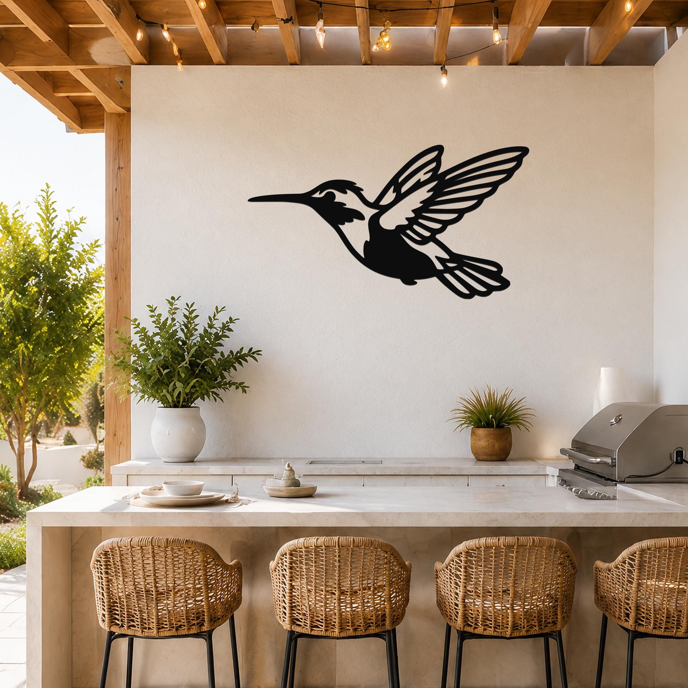 Black metal hummingbird wall art with open wings and internal cutout details, mounted on a white wall above a kitchen counter in an outdoor patio setting.