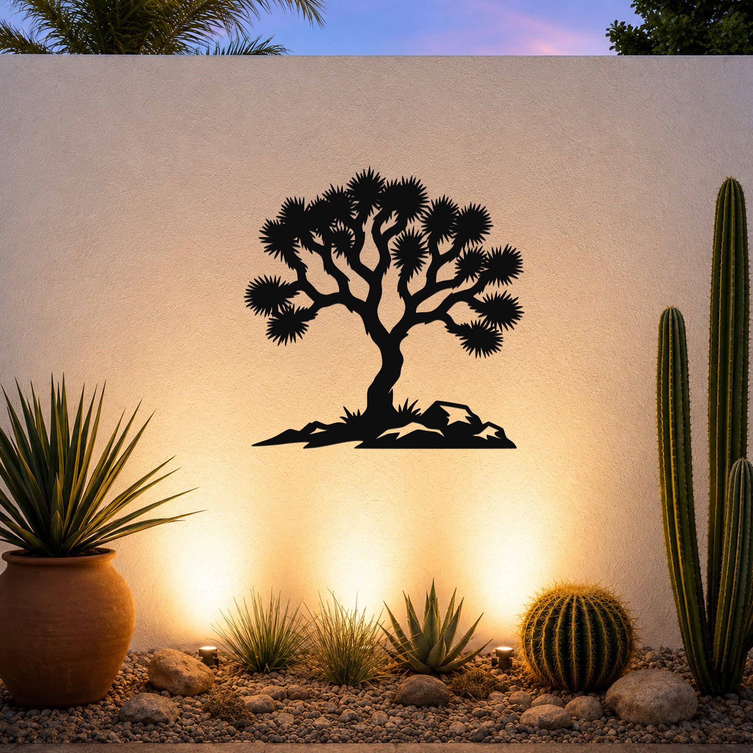 Black metal wall art silhouette of a Joshua tree with spiky branches and rocky base, mounted on an outdoor wall above a desert garden with cacti and warm uplighting.